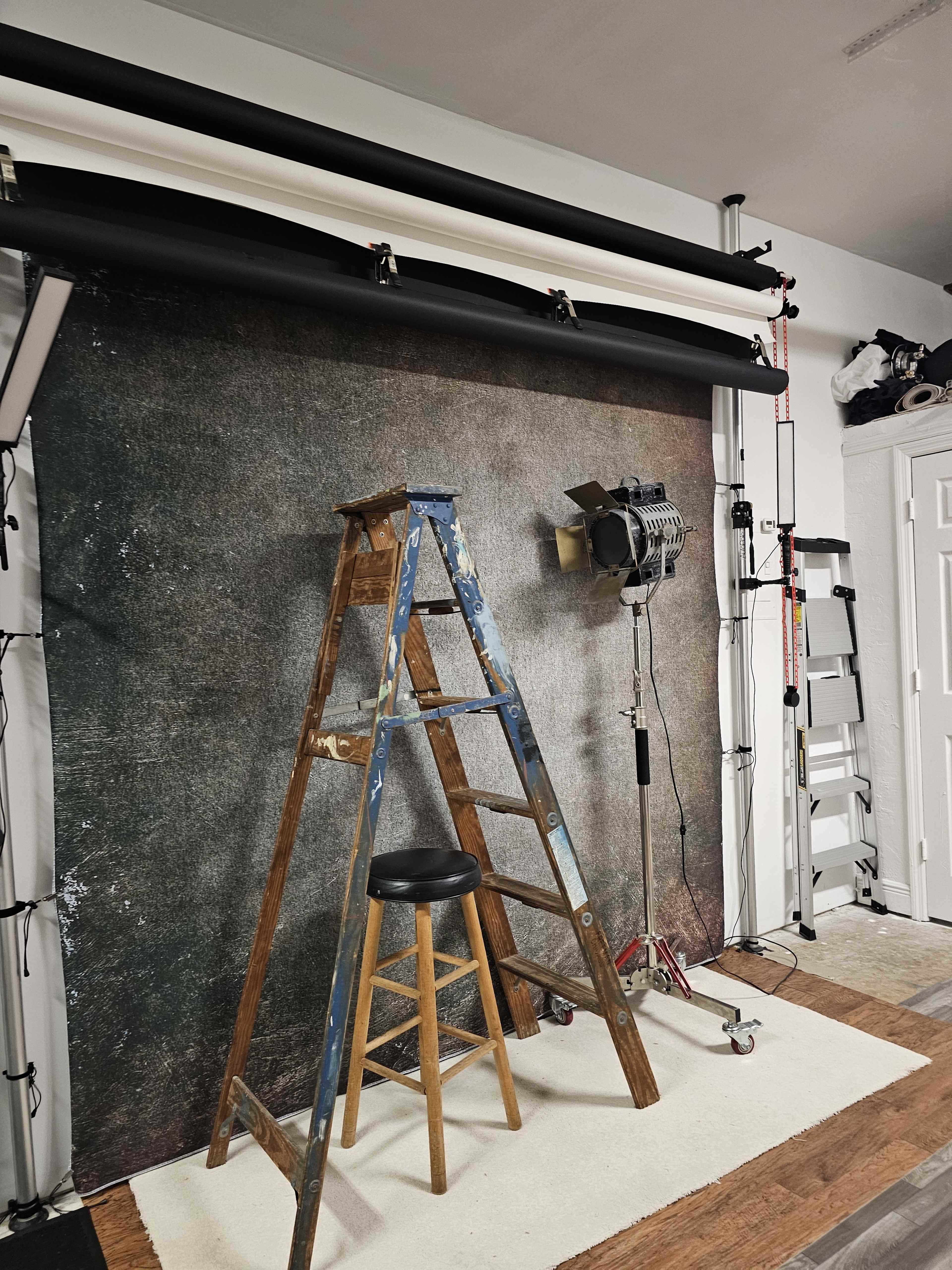 A wooden ladder and a stool are positioned in front of a textured backdrop in a studio setting, with lighting equipment nearby.