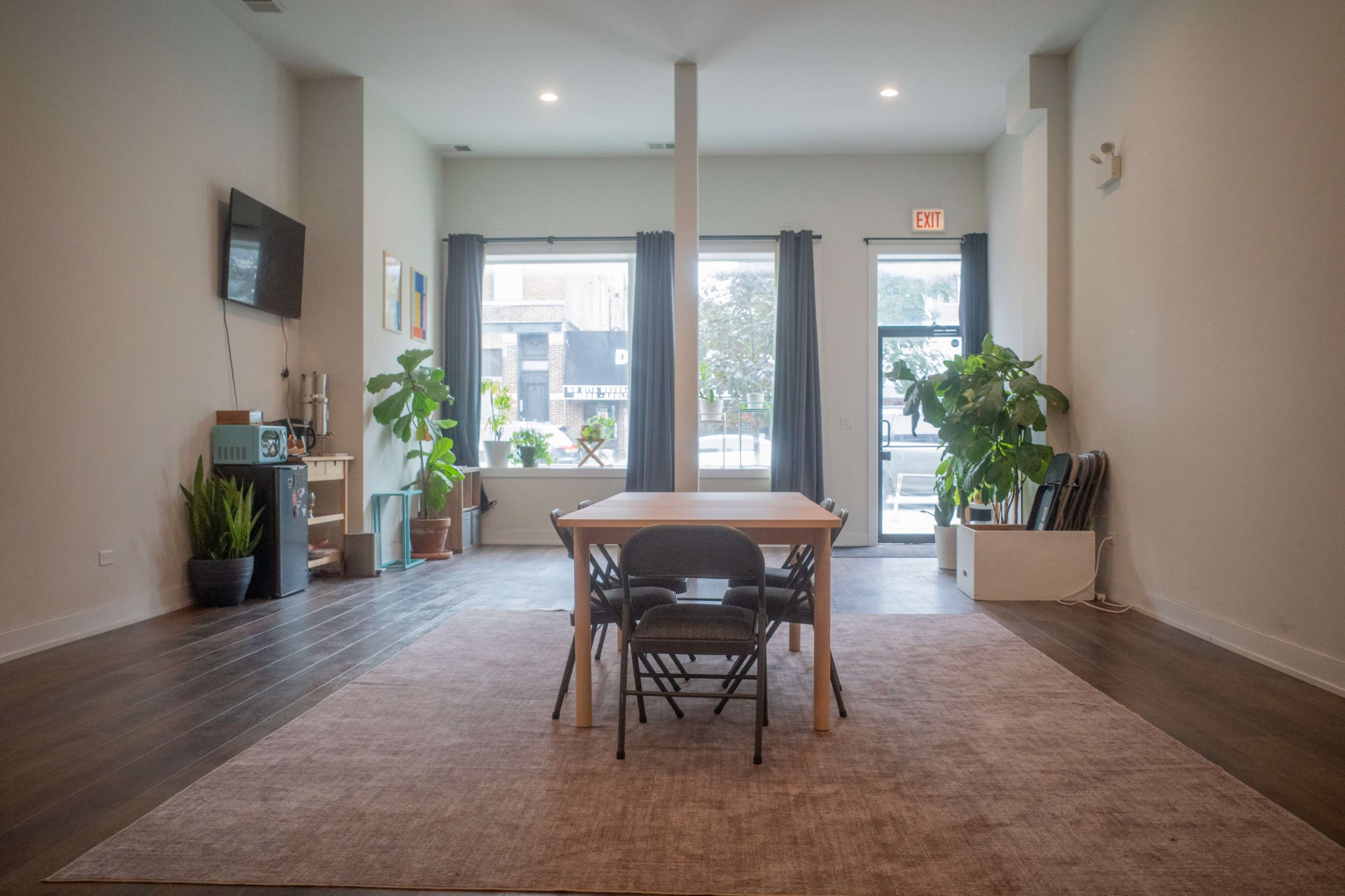 The image shows a spacious, brightly lit room with a central wooden table and chairs, surrounded by large windows and potted plants.
