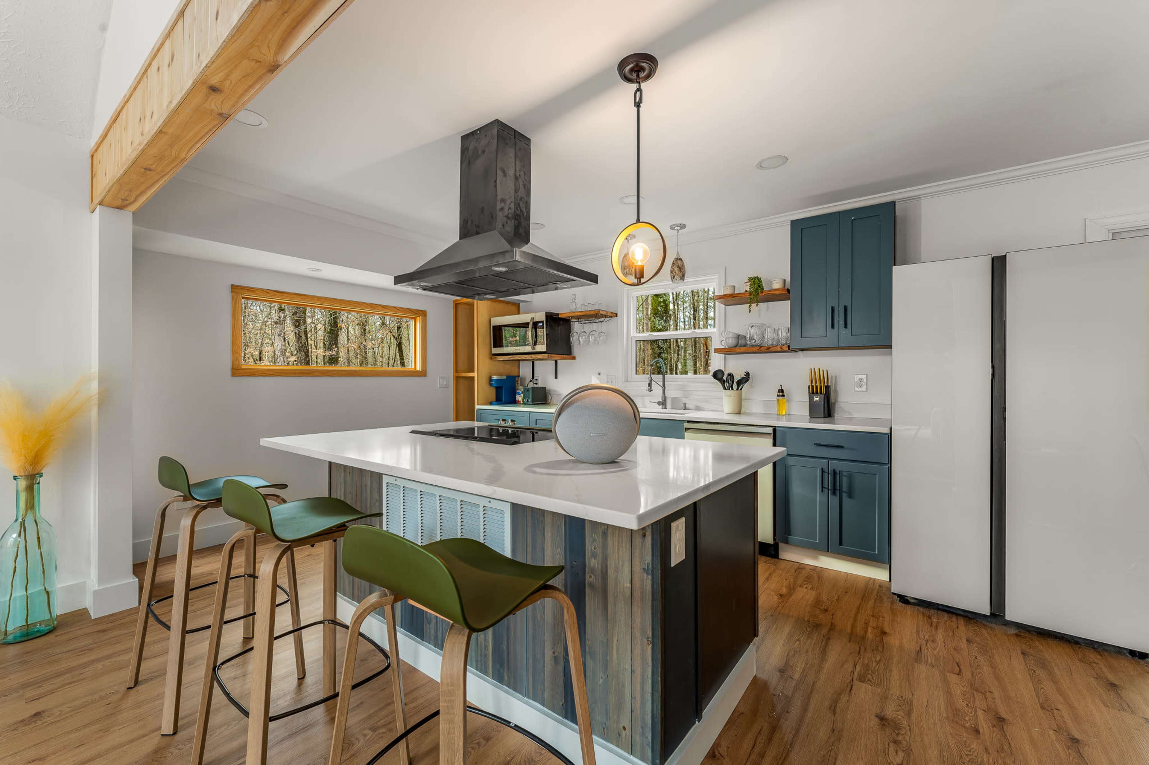The kitchen features a central island with bar seating, a metal range hood, and blue cabinets against a backdrop of large windows.