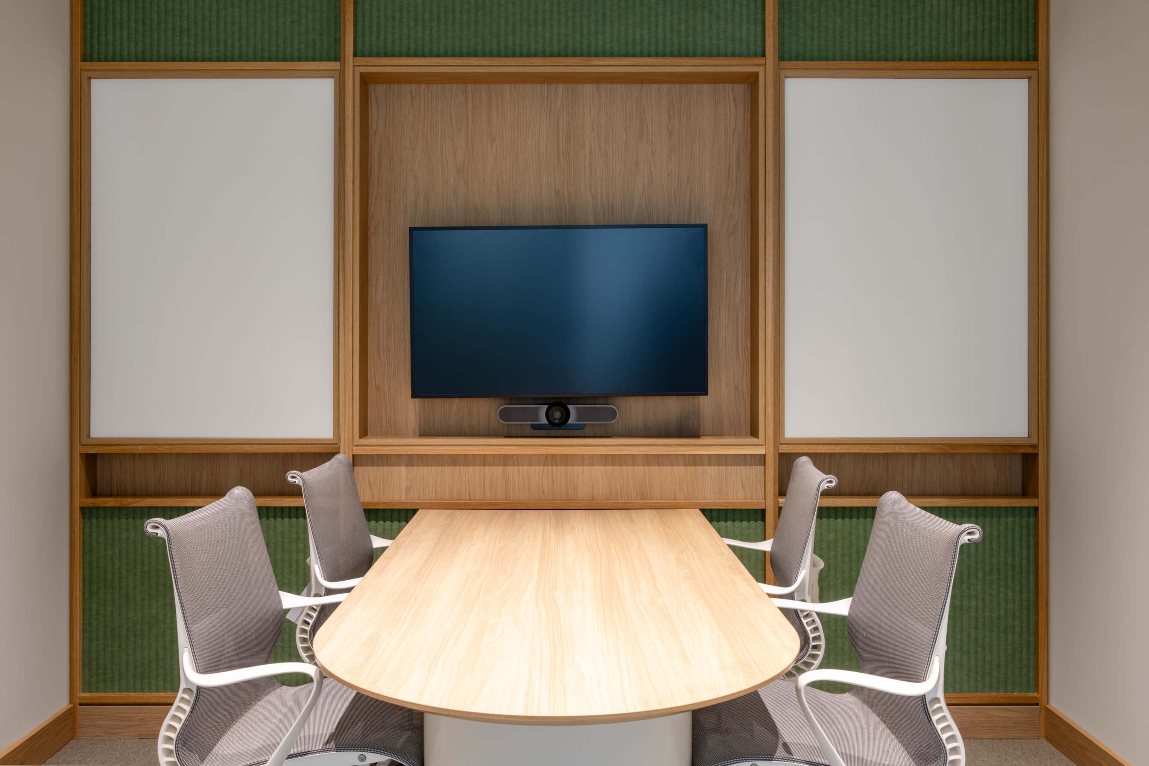 A conference room features a long wooden table surrounded by ergonomic chairs, with a television mounted on a wooden panel in the background.