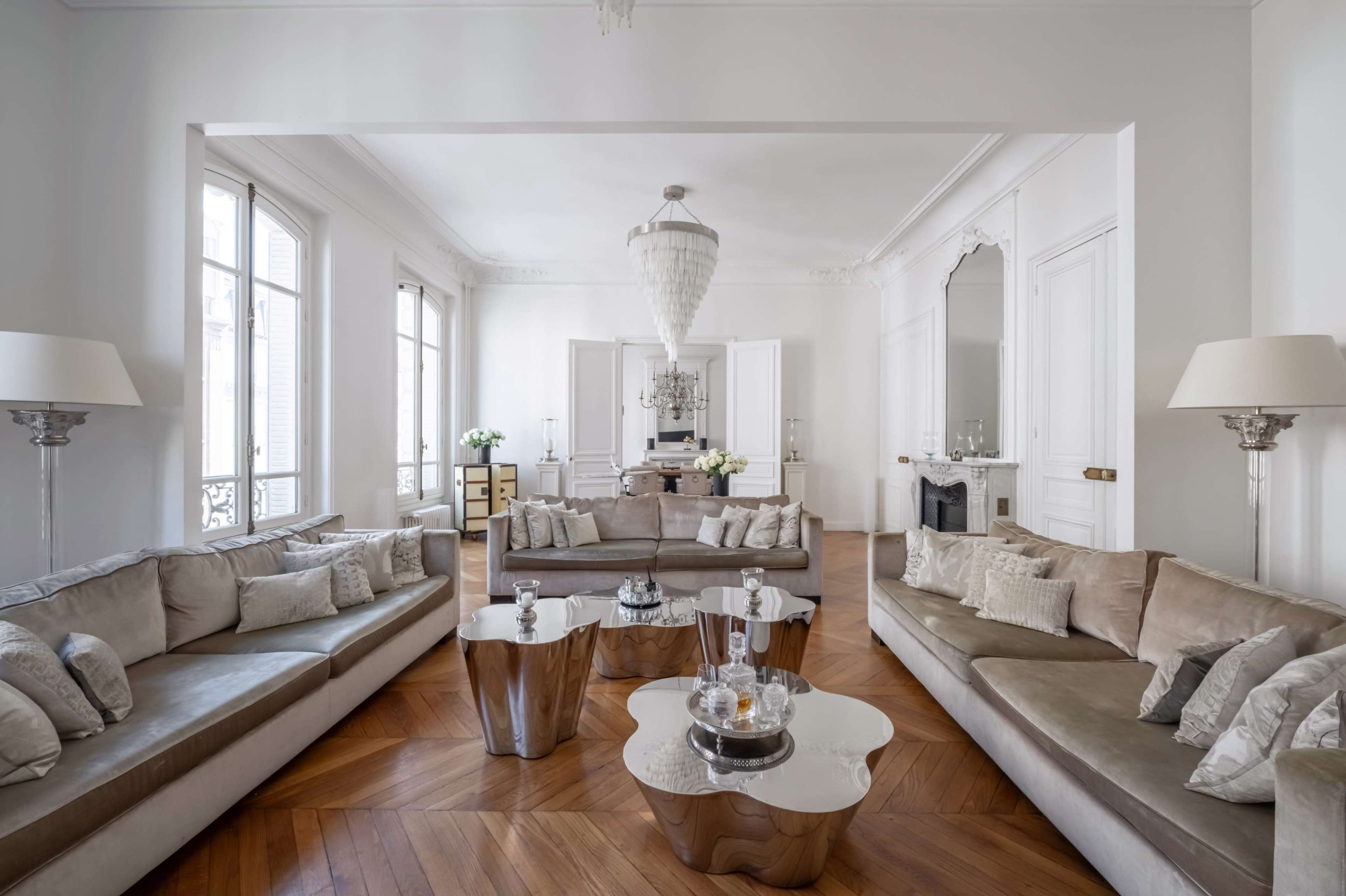 The image shows a spacious, elegant living room featuring light-colored sofas, a unique coffee table, and large windows allowing natural light to fill the space.