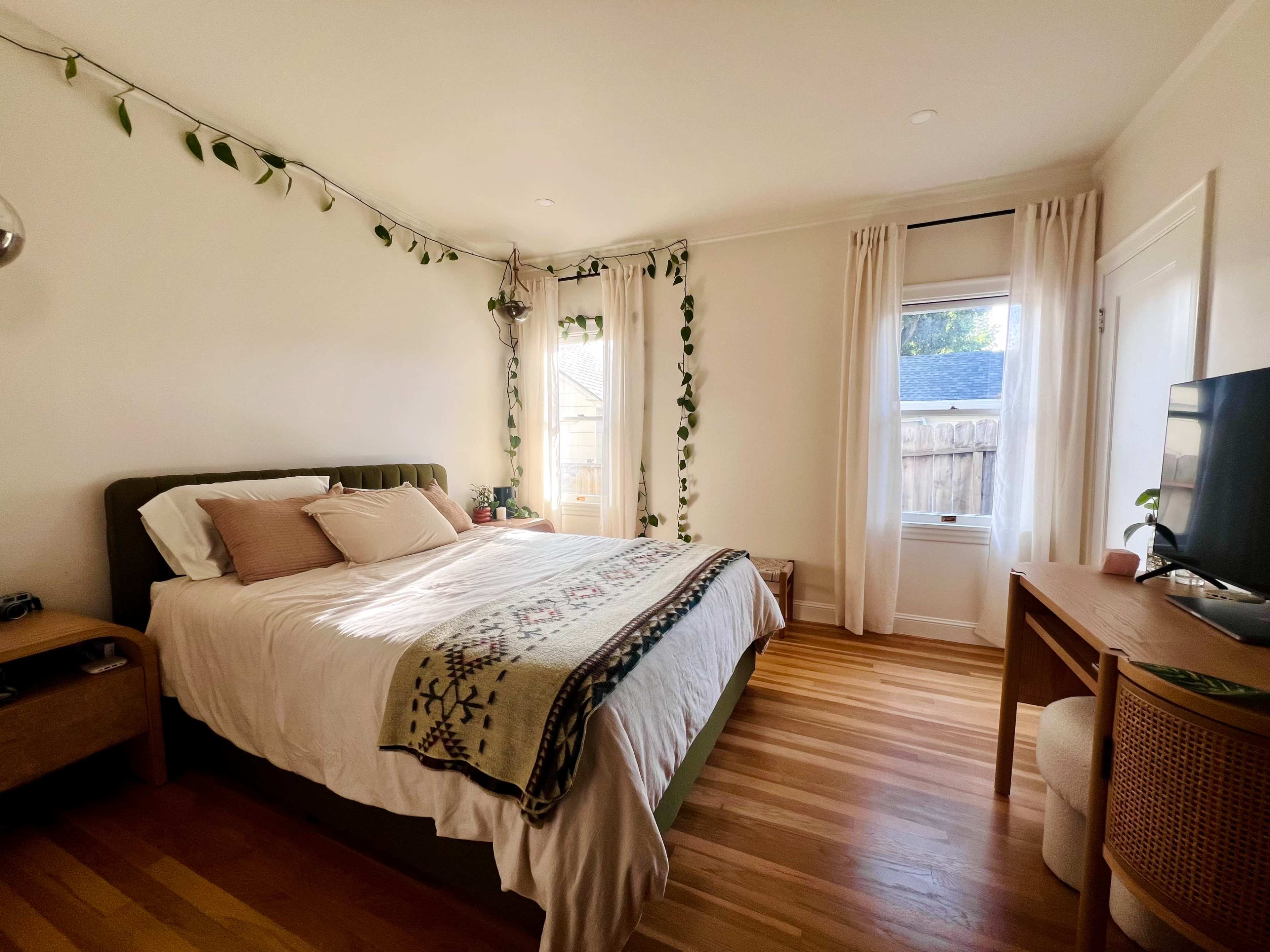 The bedroom features a bed with a patterned throw blanket, surrounded by potted plants and natural light from a window.