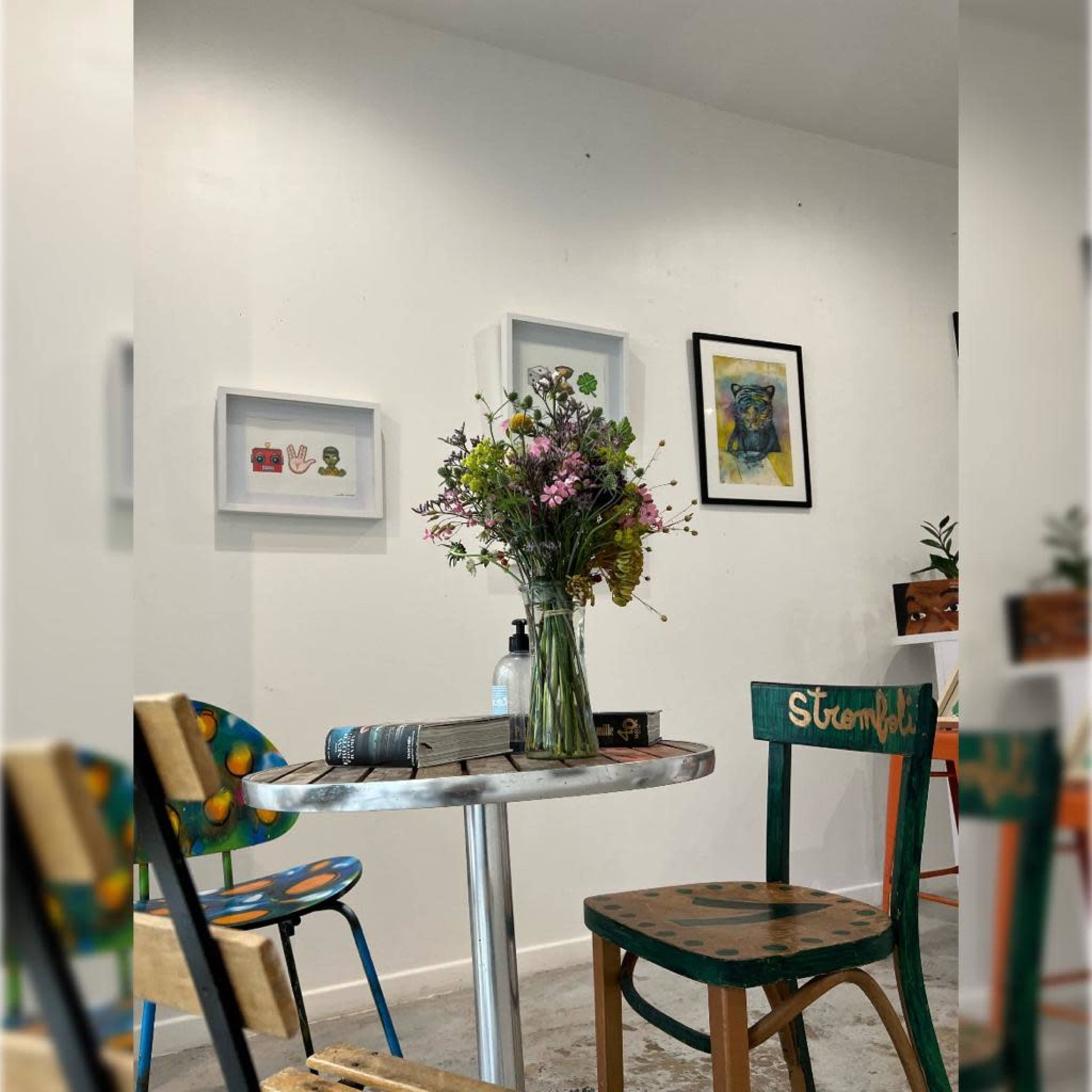 A table with a vase of flowers and an open book is positioned among colorful chairs in a gallery-like room with framed artworks on the walls.