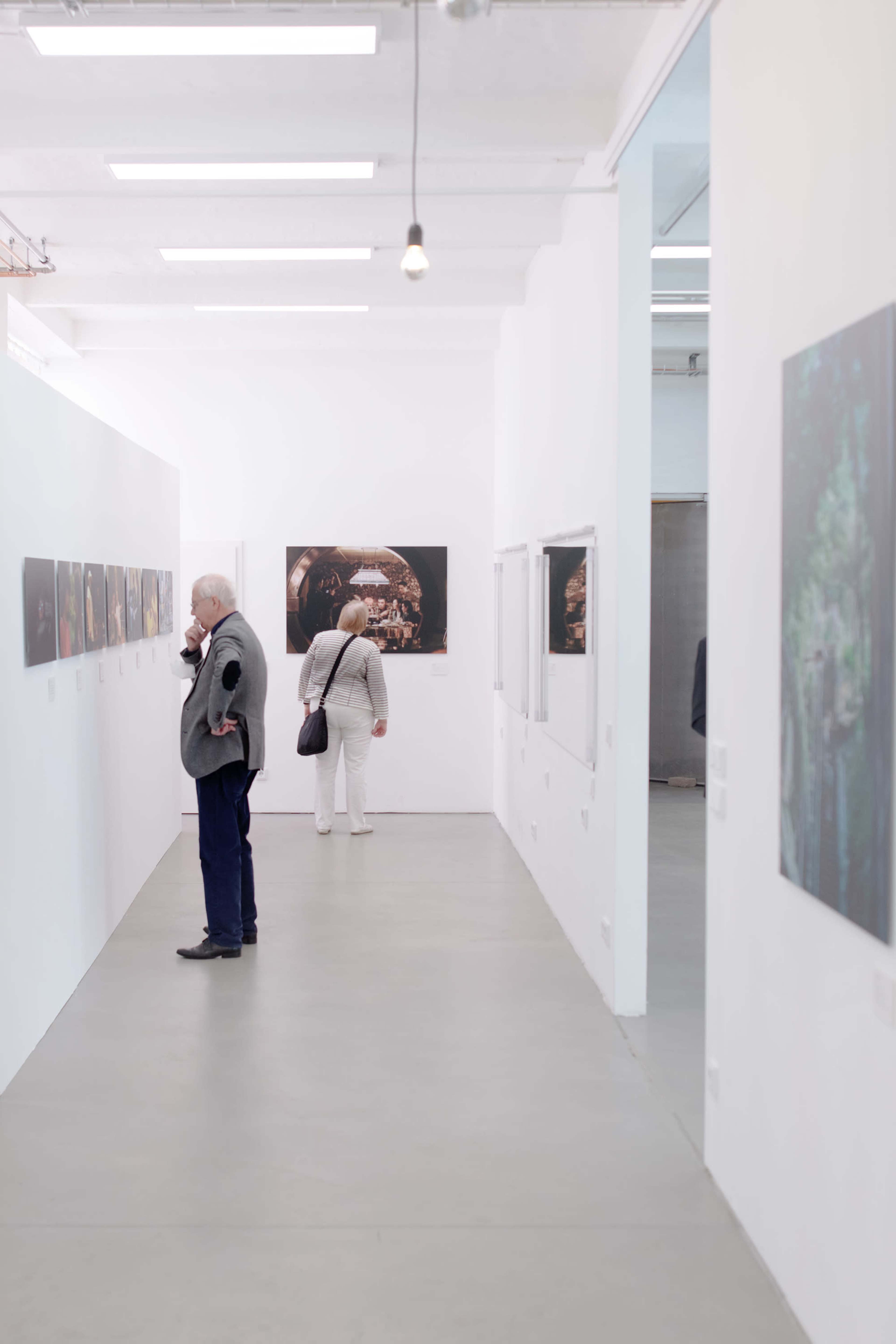 Two visitors explore an art exhibition in a bright, minimalist gallery featuring framed artworks on white walls.