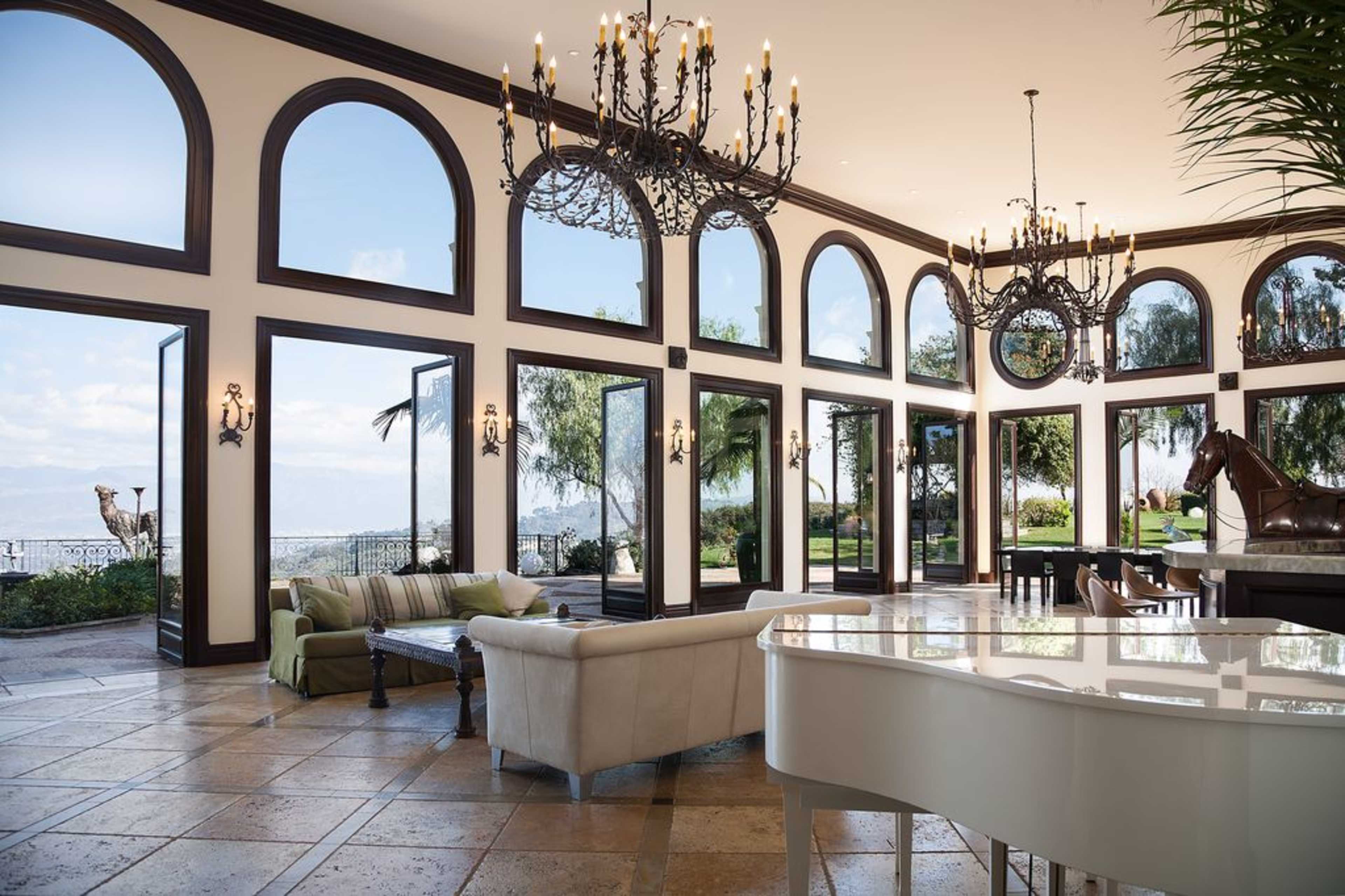 The interior of a spacious living room features large arched windows facing a scenic outdoor landscape, with a grand piano and elegant chandeliers.