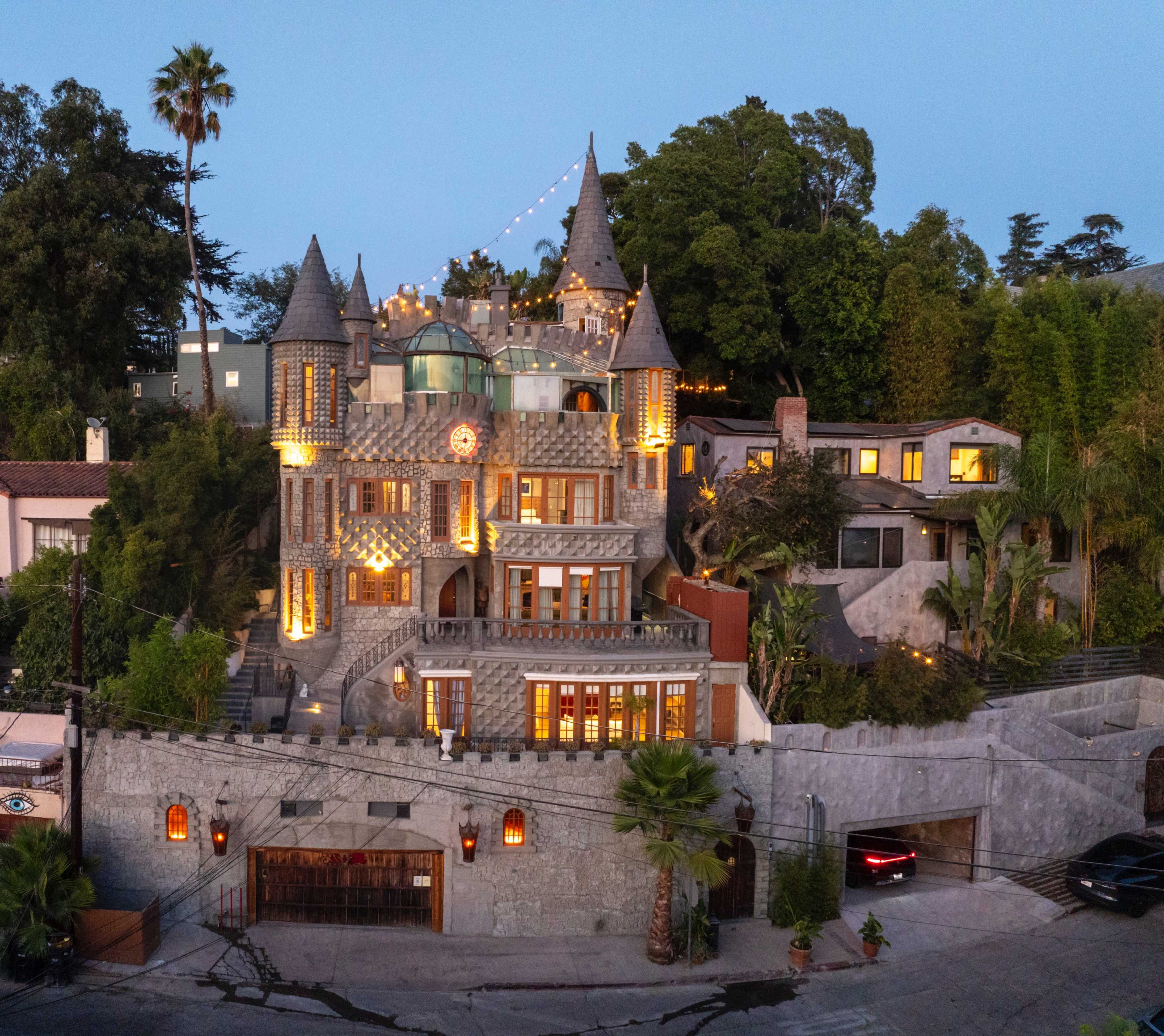 A large, intricately designed castle-style house with multiple towers and a stone exterior is illuminated in the evening, surrounded by lush trees and landscaped gardens.