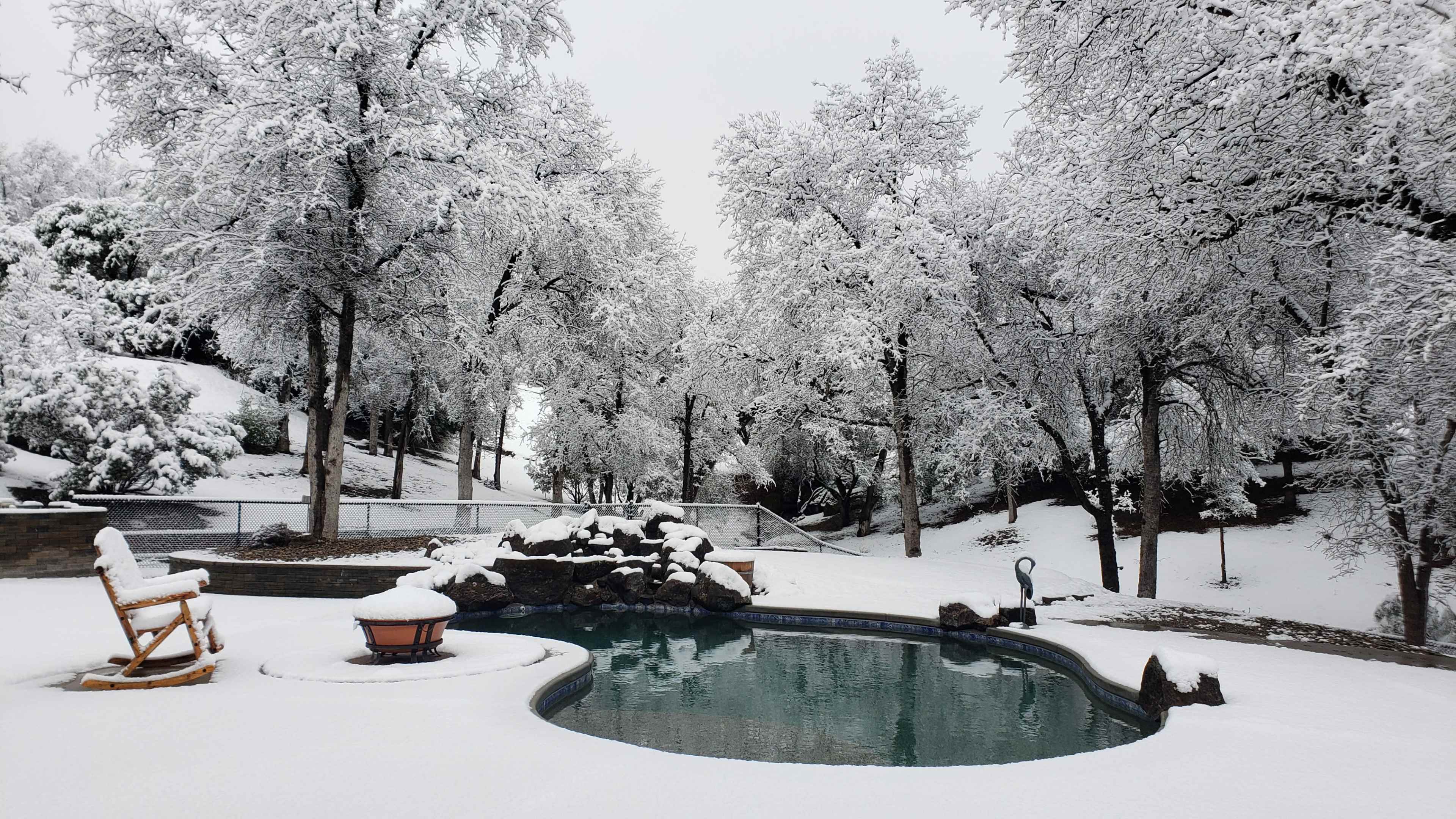 A snowy landscape surrounds a swimming pool, with frosted trees and a wooden rocking chair nearby.