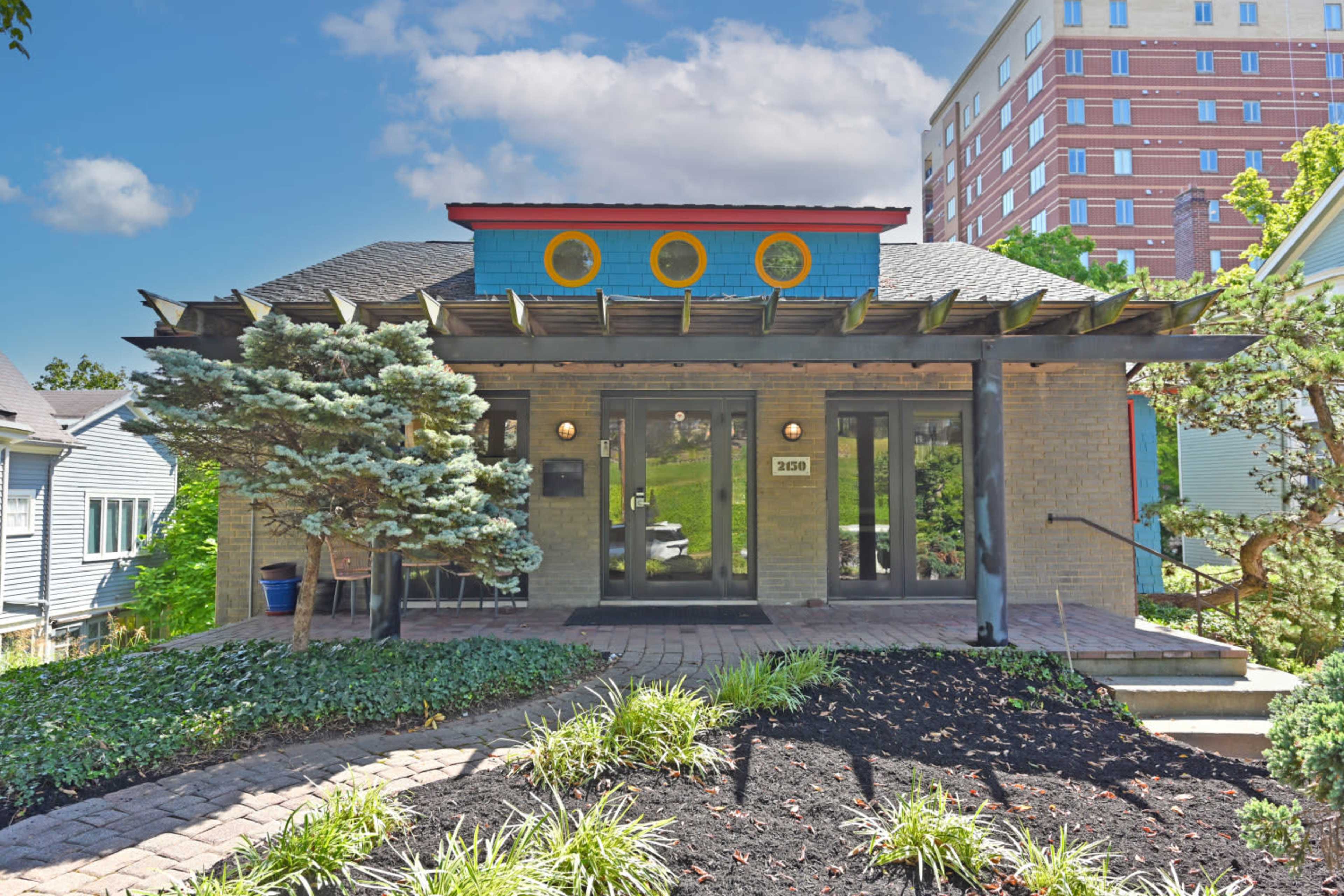 The image shows a modern-style house with a flat roof, colorful accents, and a landscaped front yard featuring greenery and stone pathways.