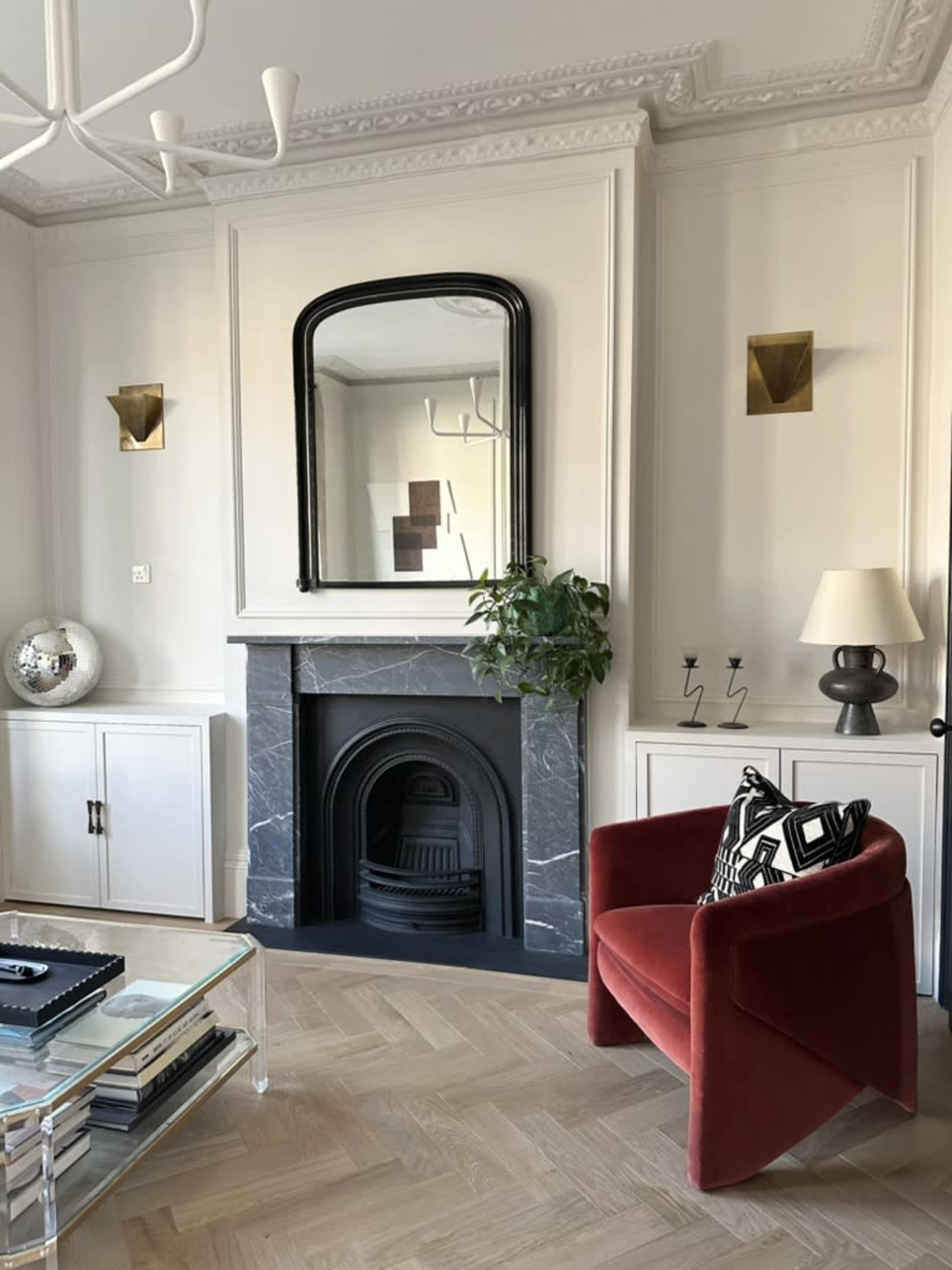 The image shows a styled living room featuring a red armchair, a marble fireplace with a mirror above it, white cabinetry, and a light fixture on the ceiling.