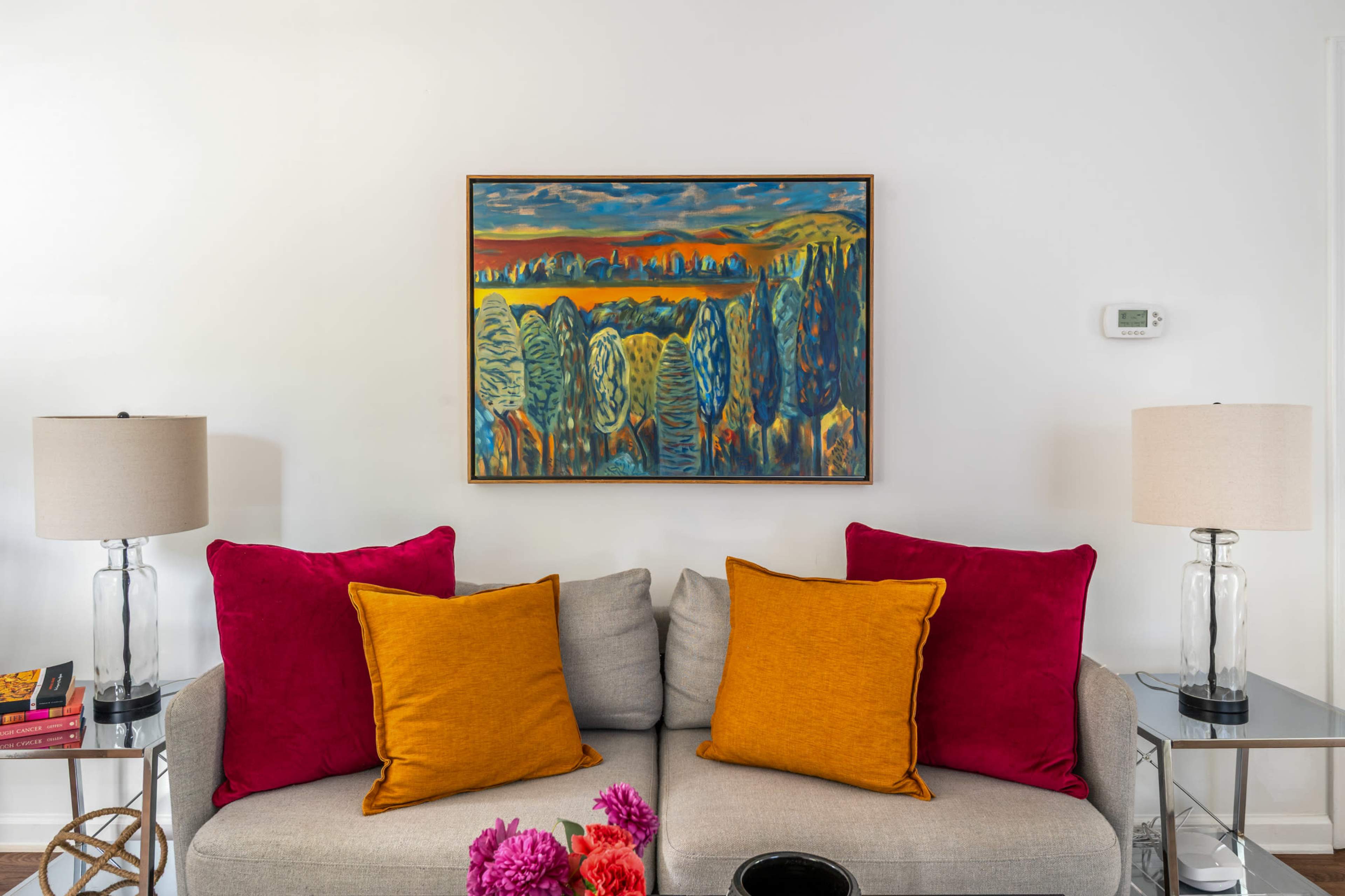 A gray sofa with orange and magenta pillows is positioned beneath a colorful landscape painting, flanked by clear table lamps on either side.