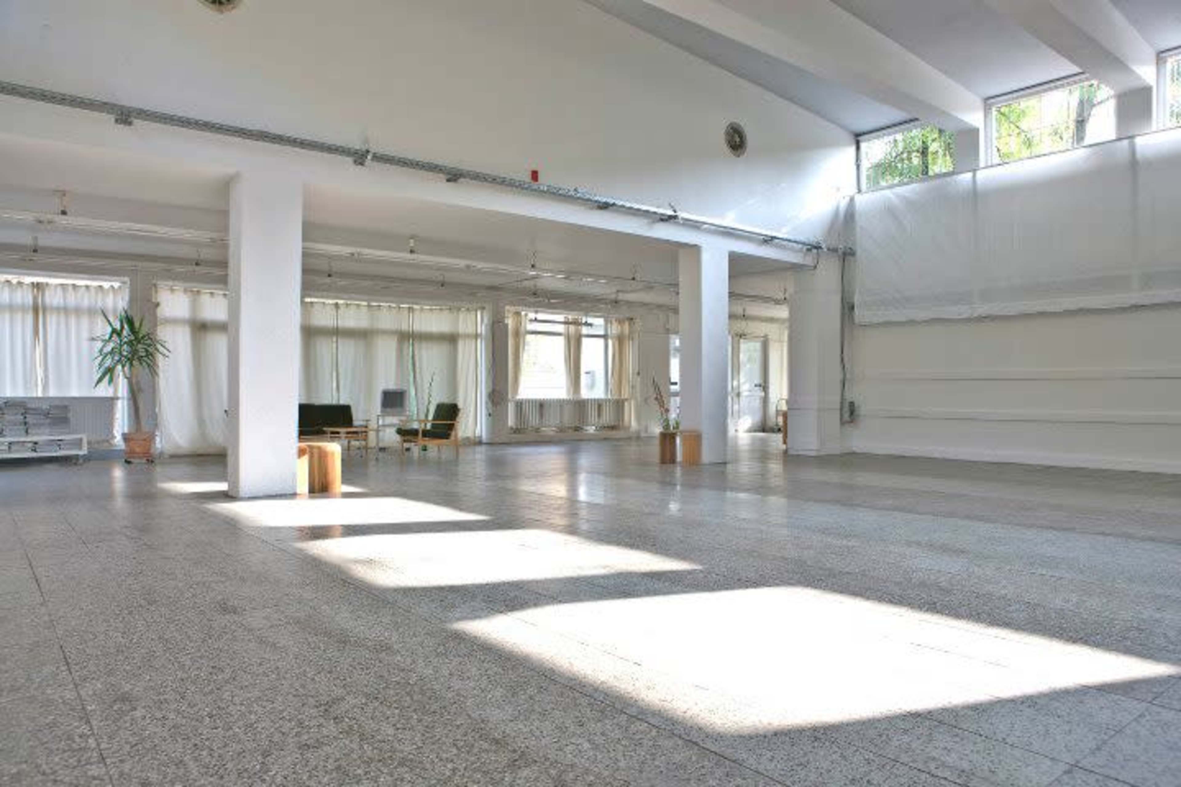 A spacious, empty room with large windows, natural light streaming in, and minimalist furniture positioned near the walls.