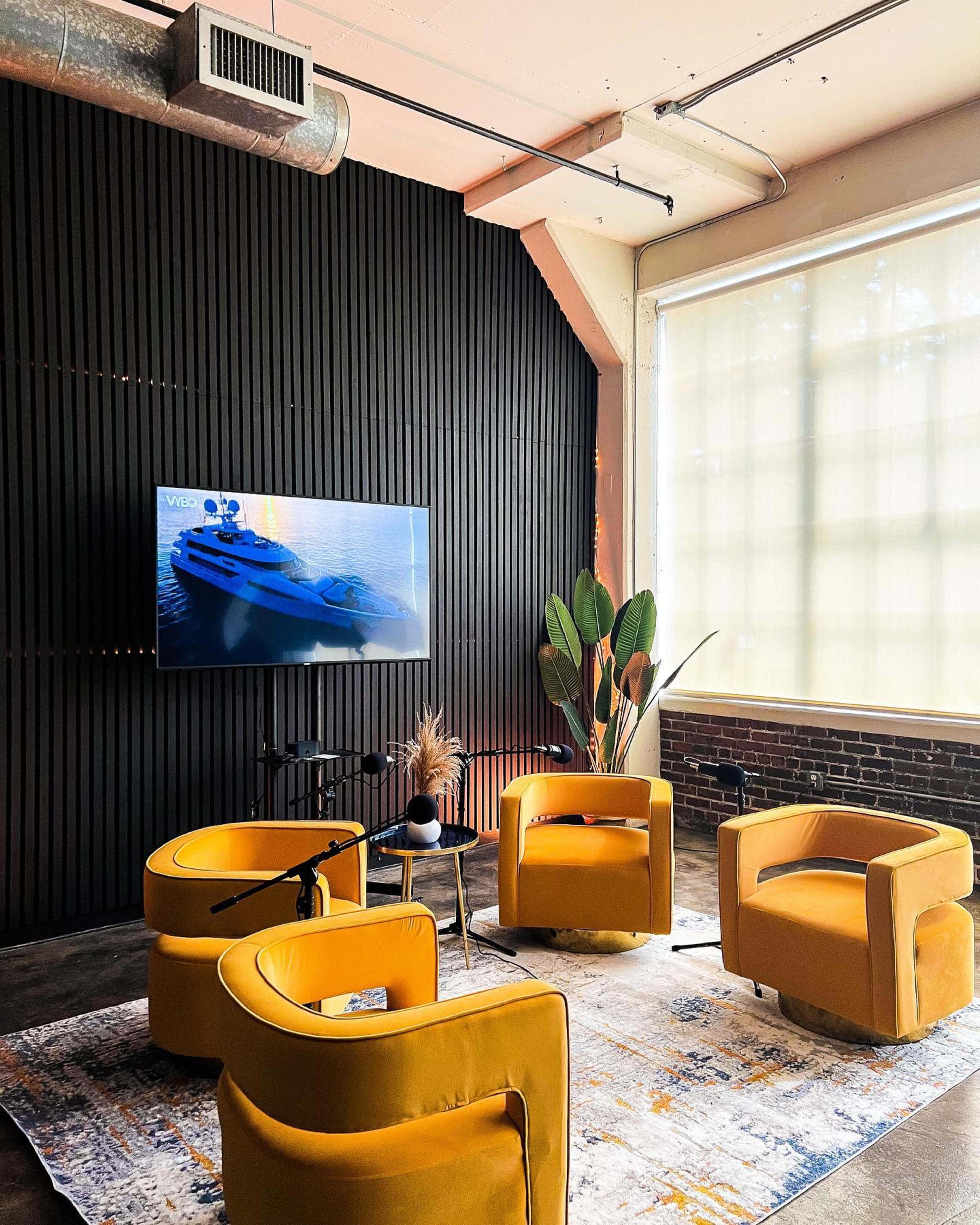 The image depicts a cozy lounge area featuring four yellow chairs arranged around a decorative rug, with a television mounted on a wooden wall and large windows allowing natural light.