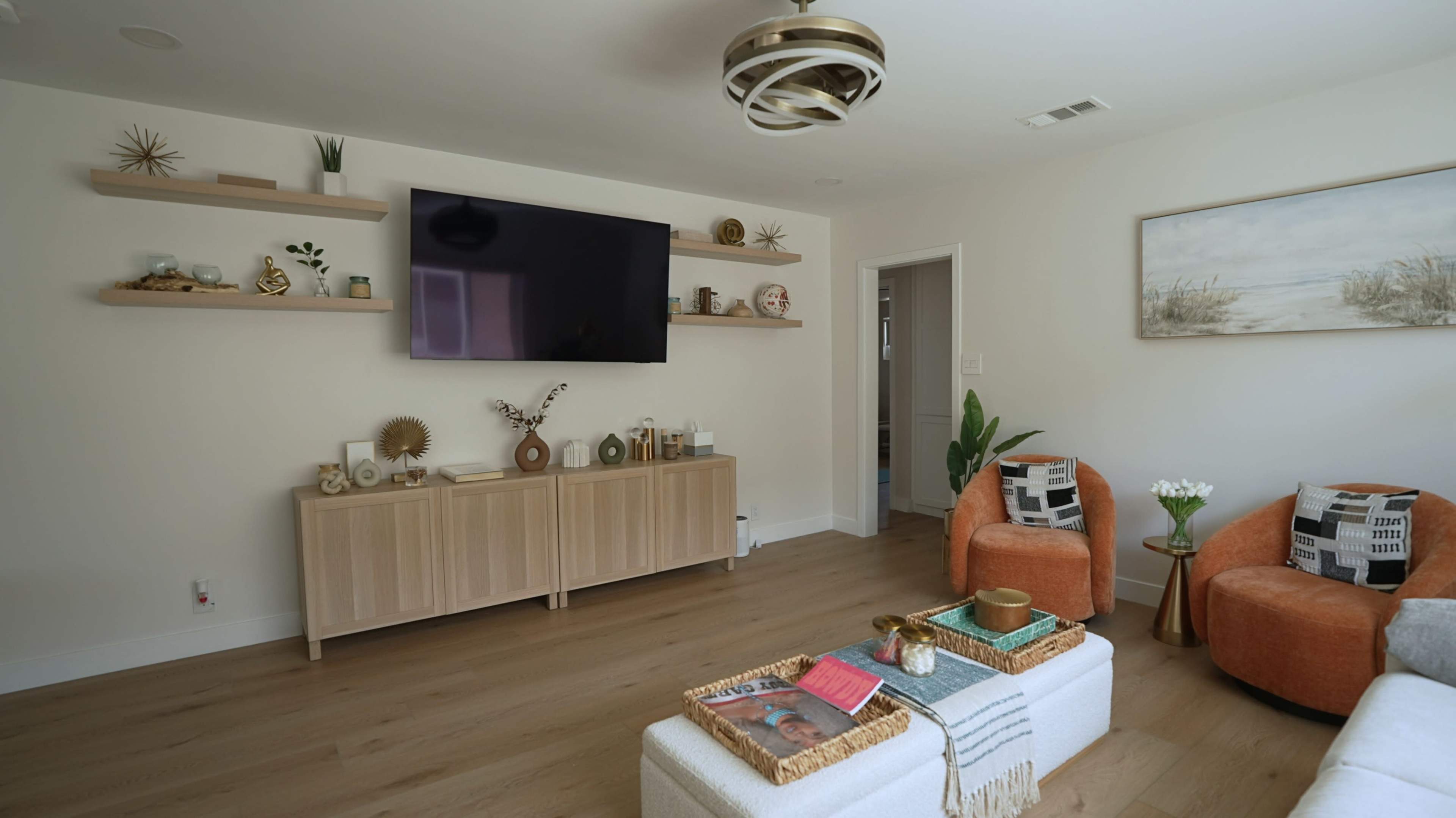 A modern living room features a large wall-mounted television above a light wooden cabinet, surrounded by decorative shelves, with an orange accent chair and a coffee table in the center.