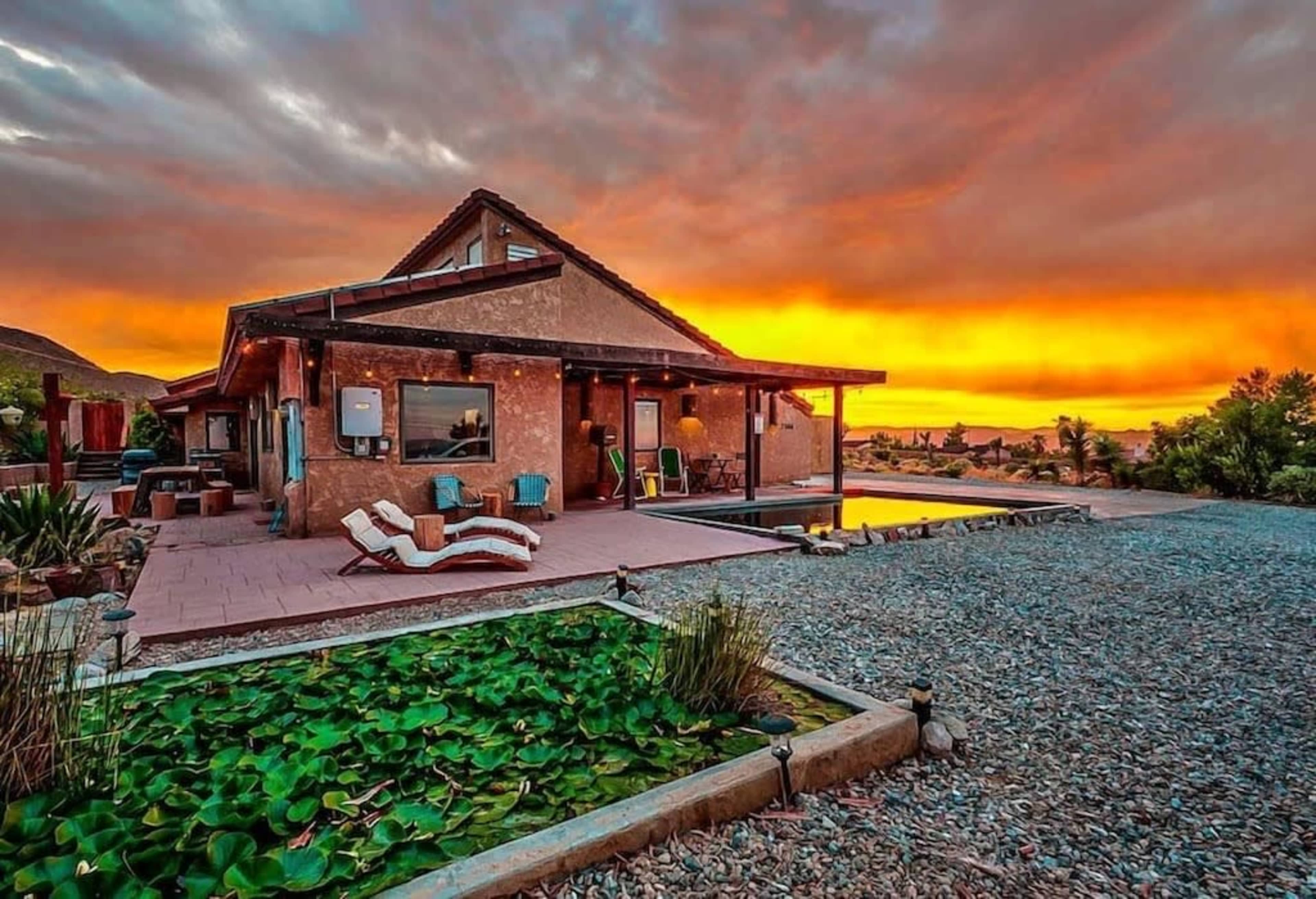 A modern house with a patio and pool is set against a vibrant sunset sky, surrounded by gravel landscaping and water lilies.