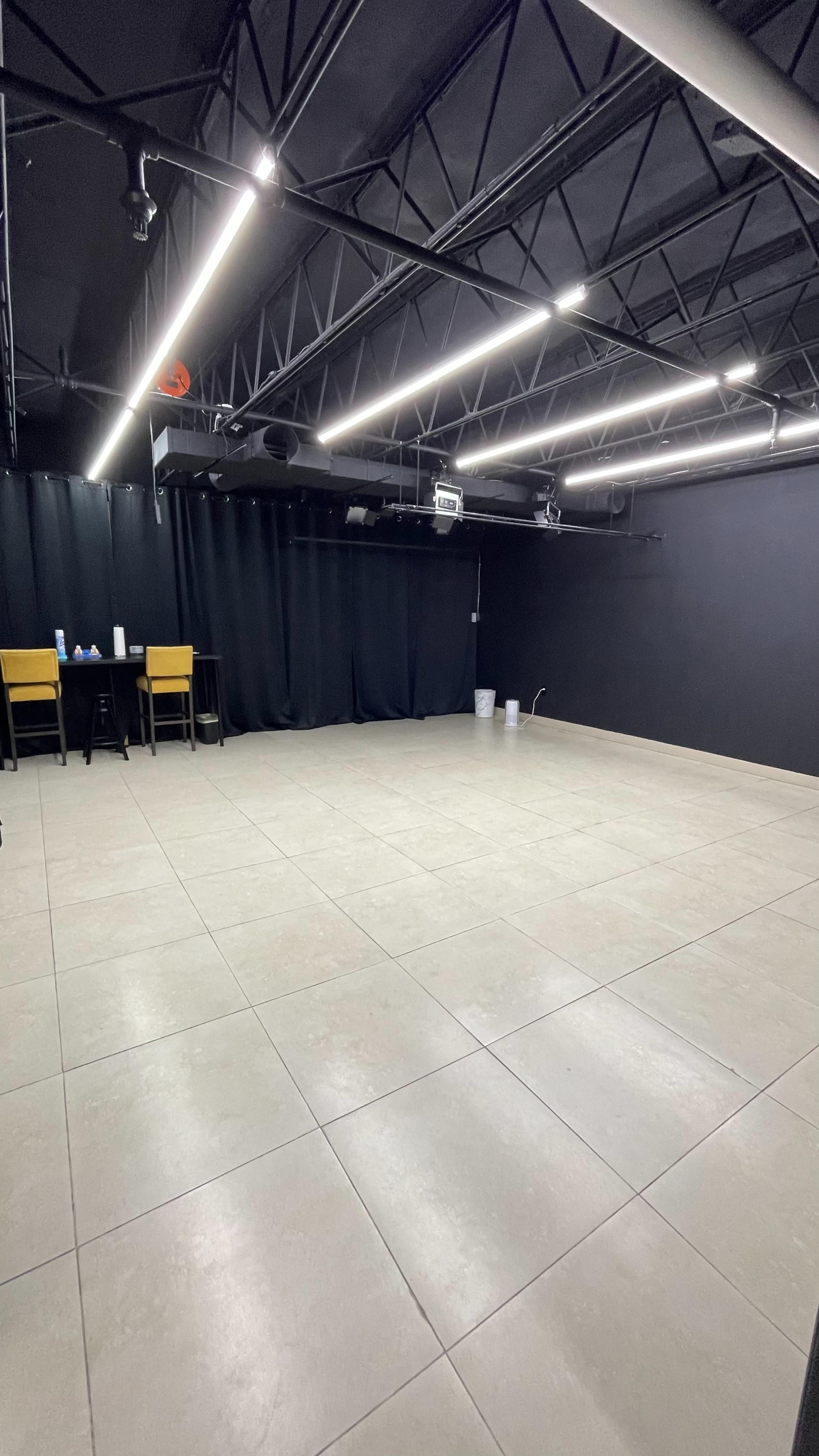 The image shows a spacious, empty room with a black ceiling and walls, featuring tiled flooring and simple furniture along one side.