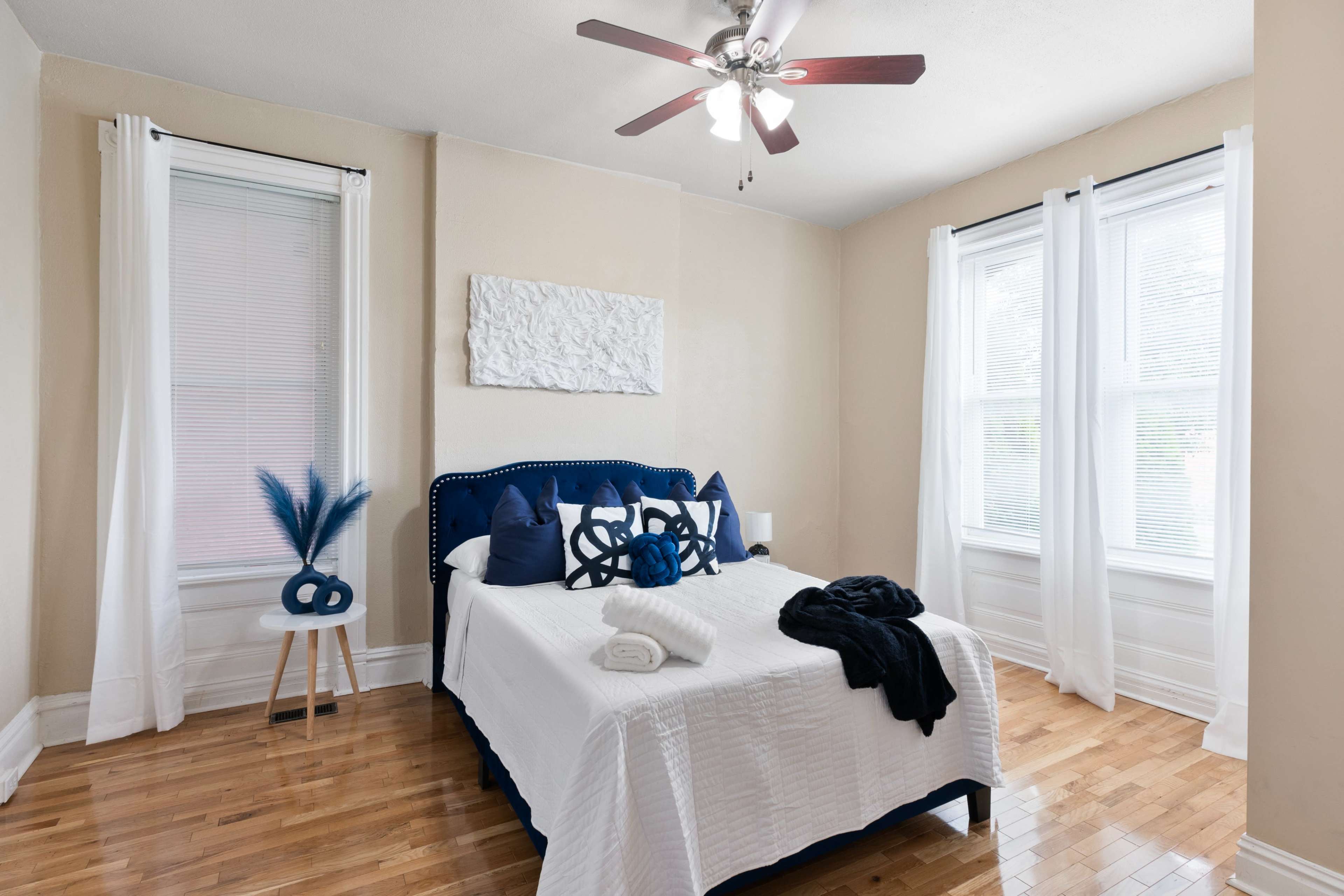 A neatly arranged bedroom featuring a blue upholstered bed, white bedding, and wooden flooring, with two windows allowing natural light to enter.