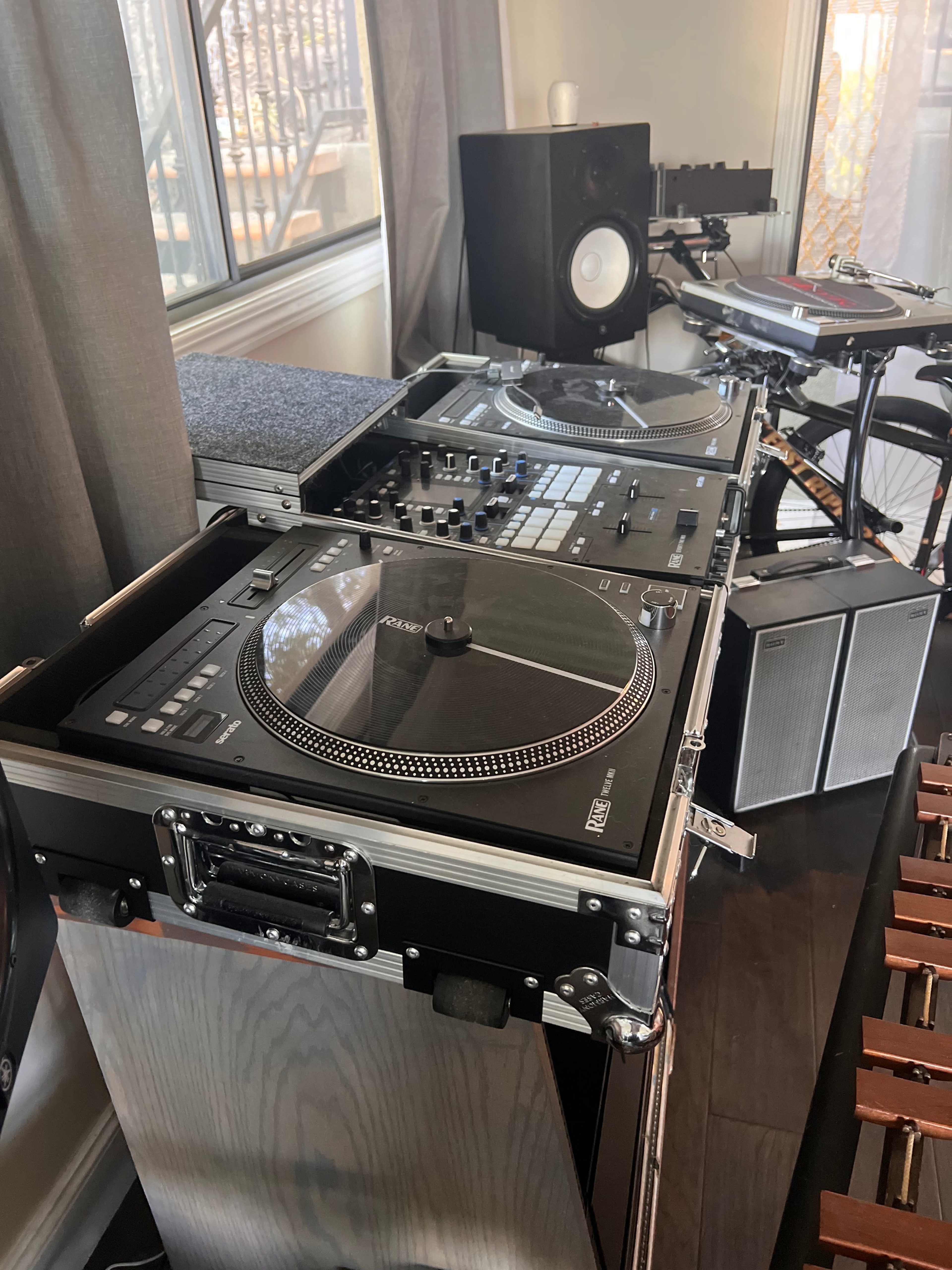 A mixing console and turntables are set up on a table next to speakers and a xylophone in a well-lit room.