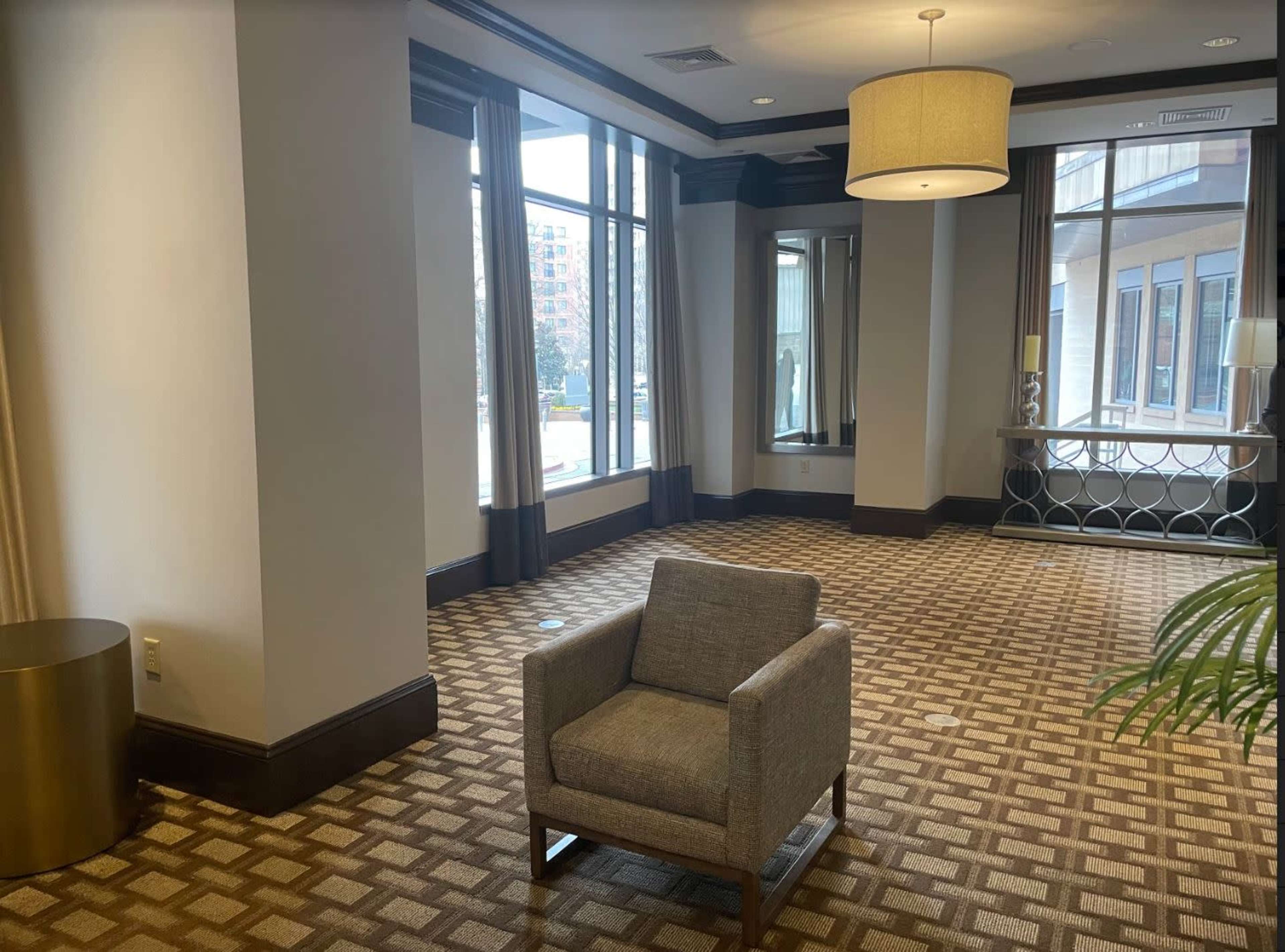 A well-lit hotel lobby area featuring a single gray armchair, a round light fixture, and large windows with views outside.