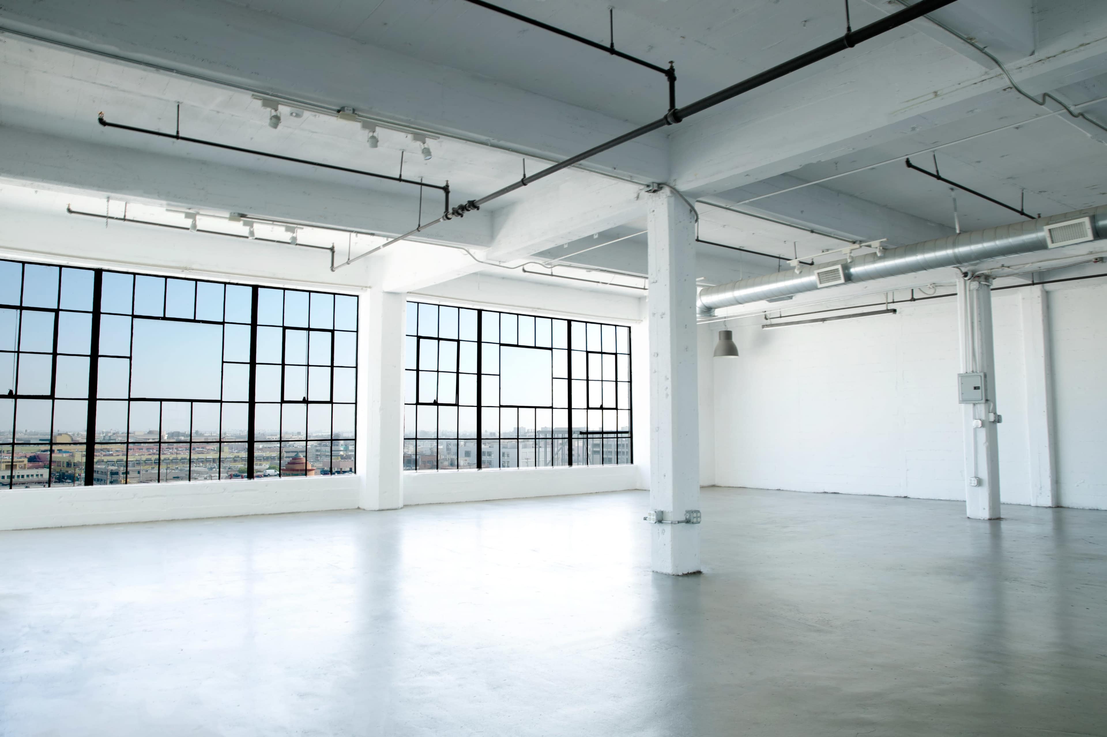 The image shows a large, empty industrial space with concrete flooring and expansive windows providing a view of the outside.
