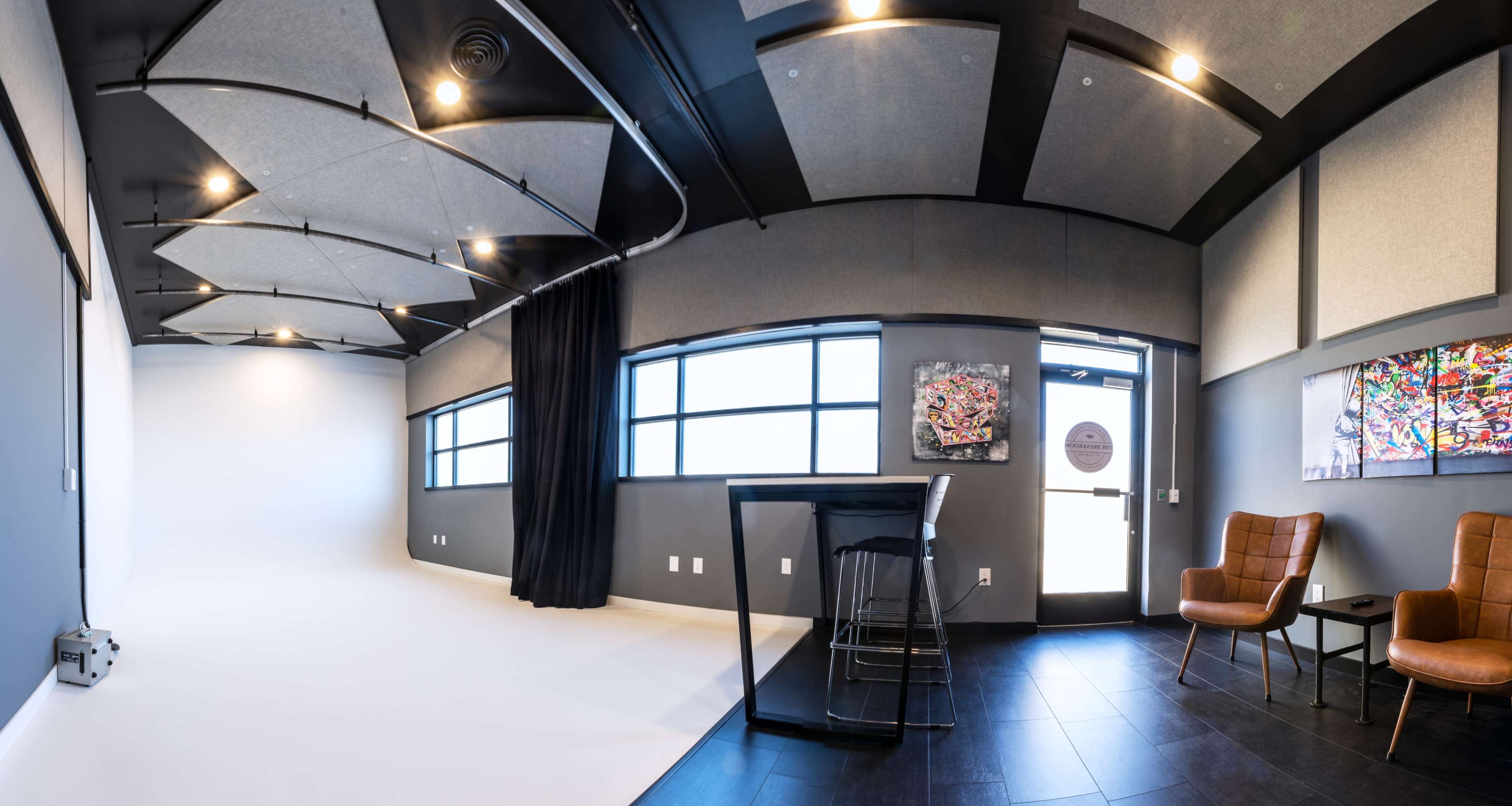 A modern studio space with a white backdrop, black curtains, a counter with stools, and two brown chairs near a door displaying artwork on the walls.