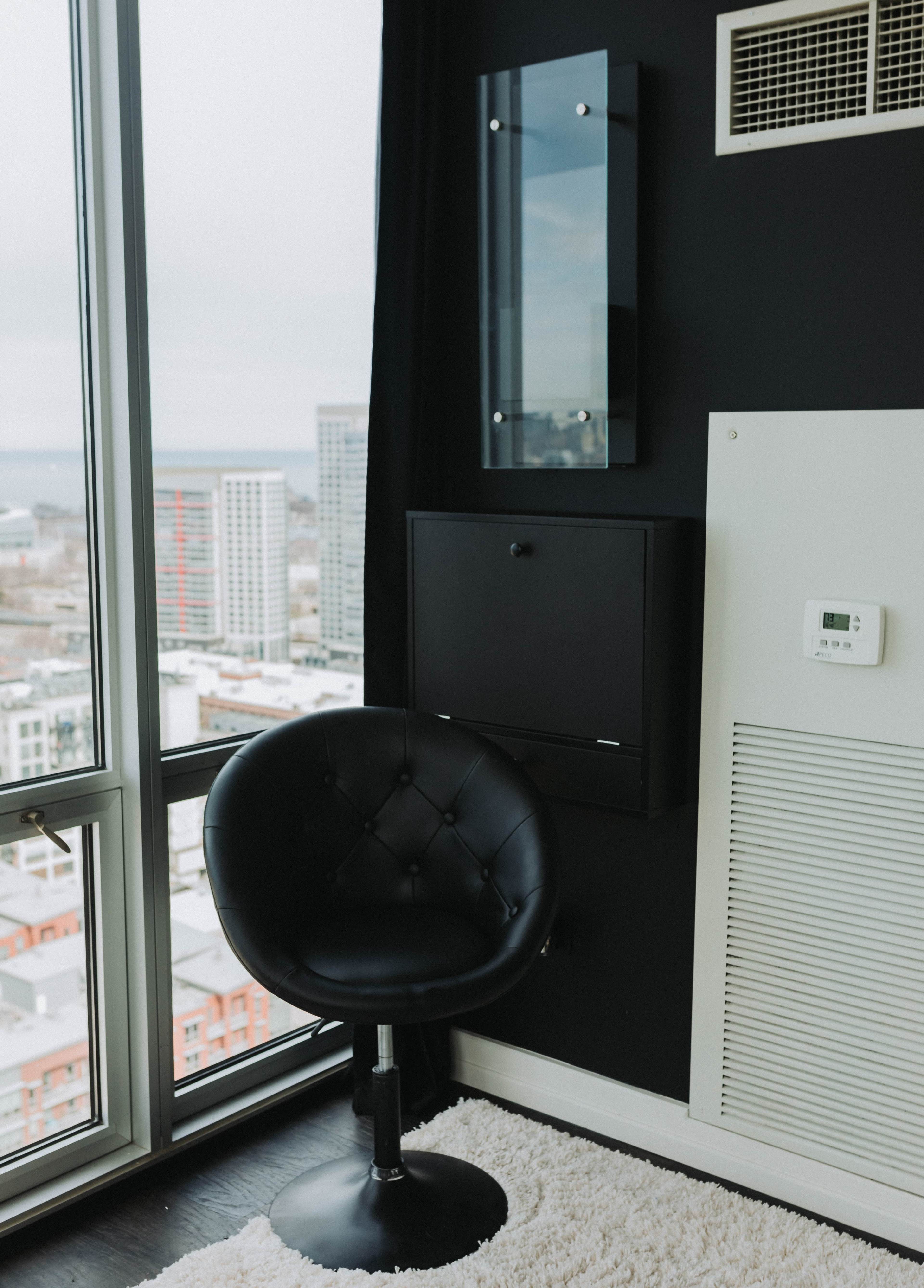 A modern black chair sits on a rug in a corner with large windows showcasing a cityscape.