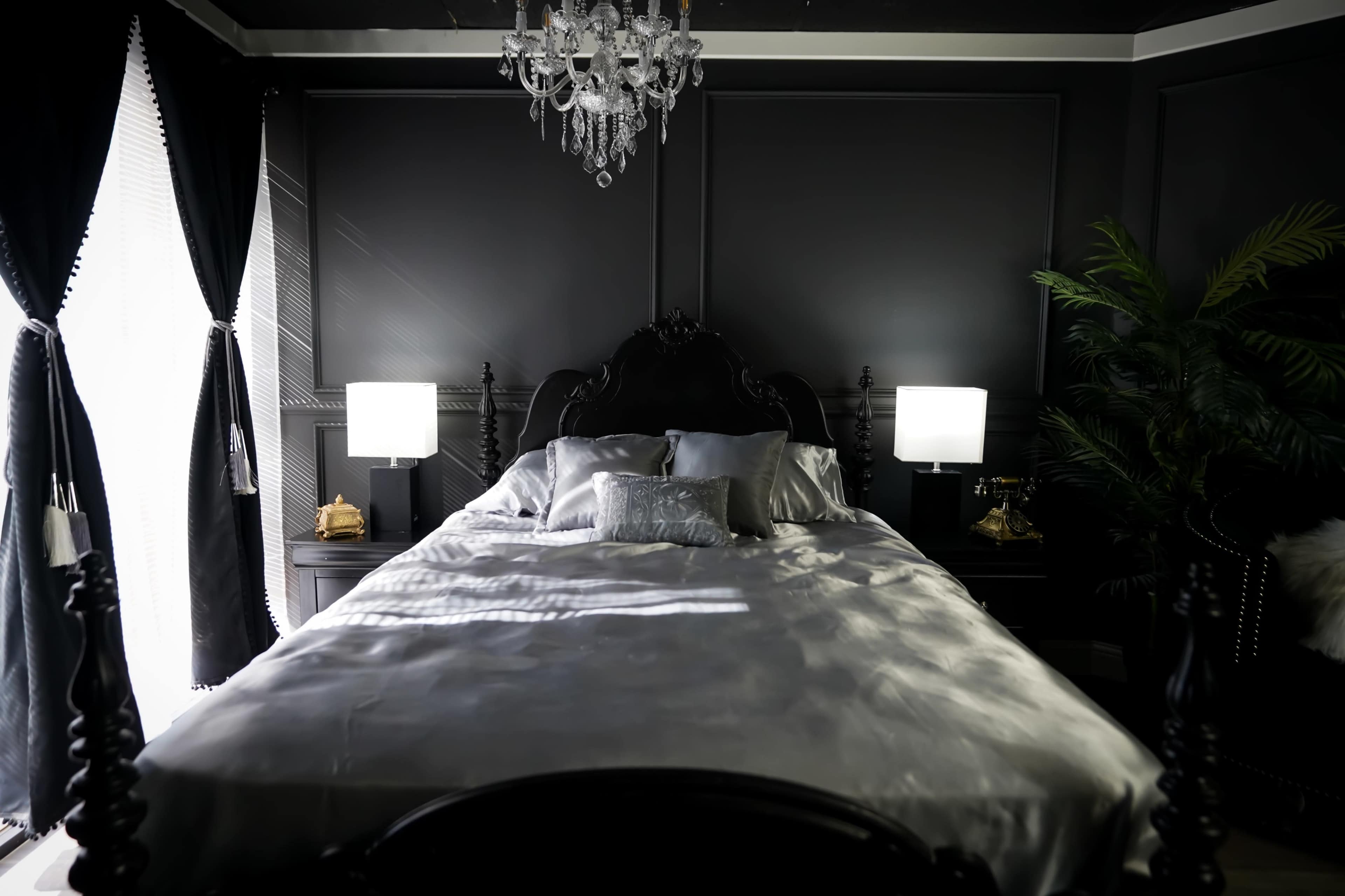 A dark, elegantly furnished bedroom features a large bed with silver bedding, flanked by two white lamps, and a chandelier overhead.