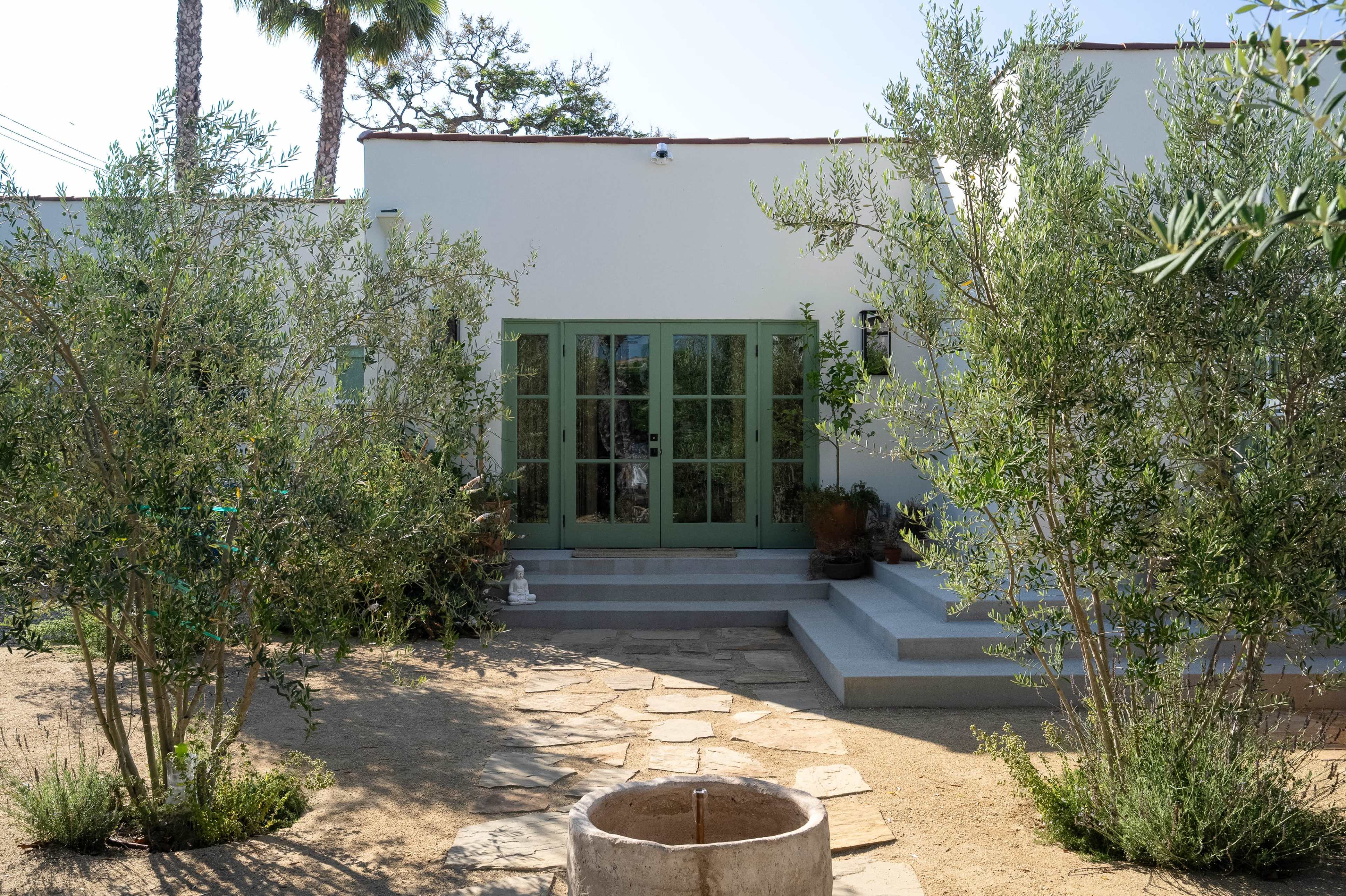 The image depicts a modern-style house with green French doors, surrounded by olive trees and a stone pathway leading to a circular stone well.