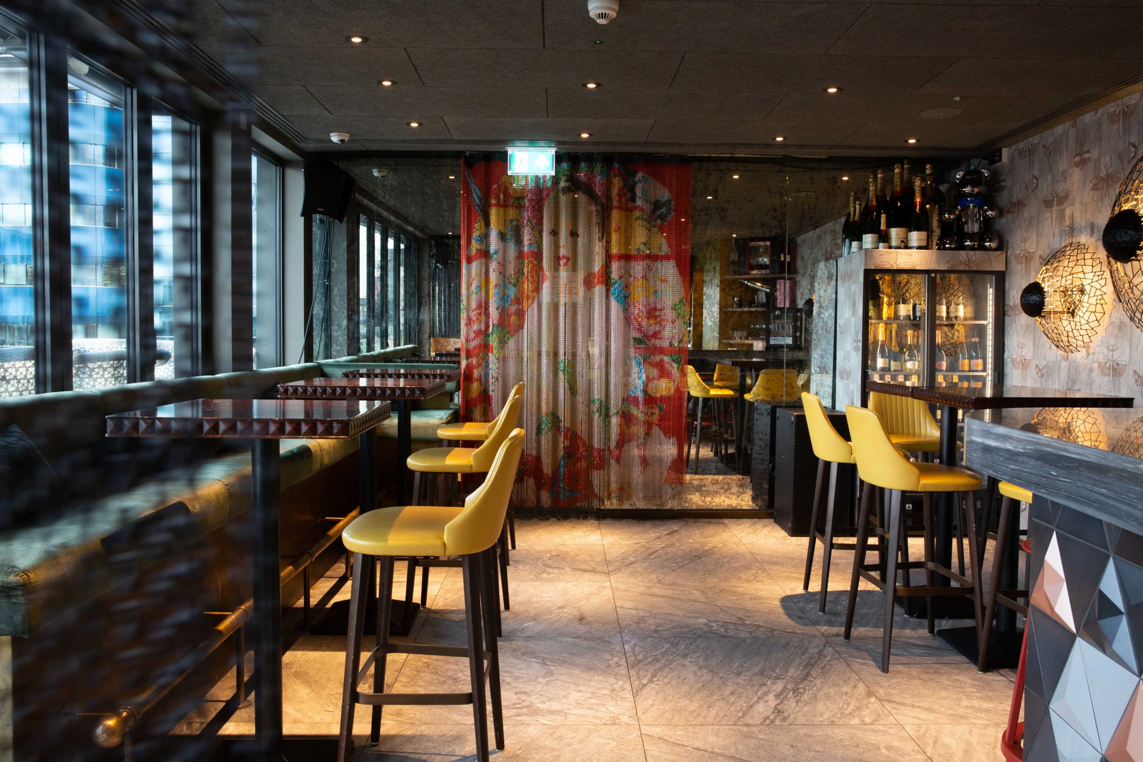 The image shows a modern bar setting with yellow stools, a decorative floral curtain, and shelves stocked with bottles.