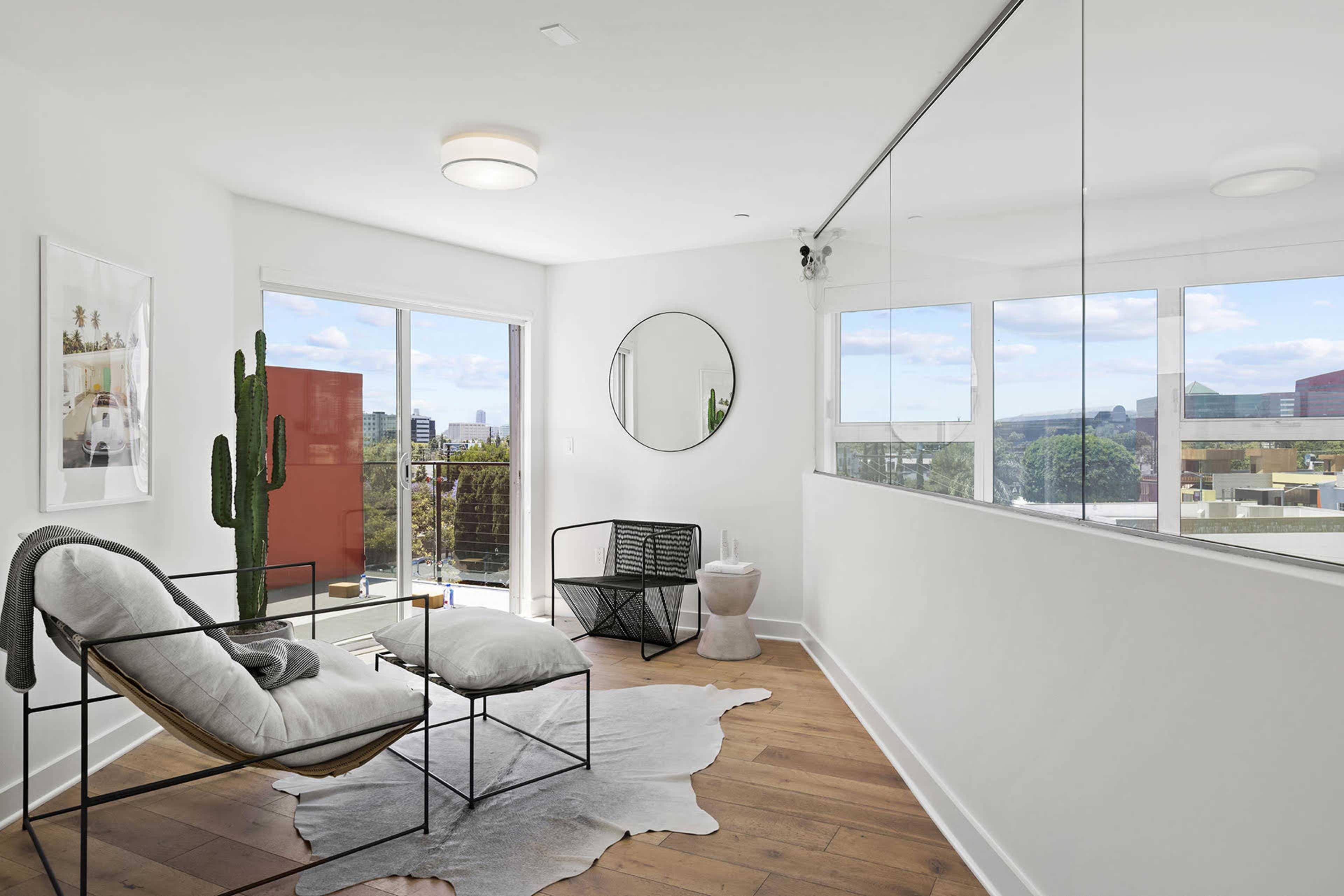 A bright room features a chair, a small table, a cactus, and large windows with a view.