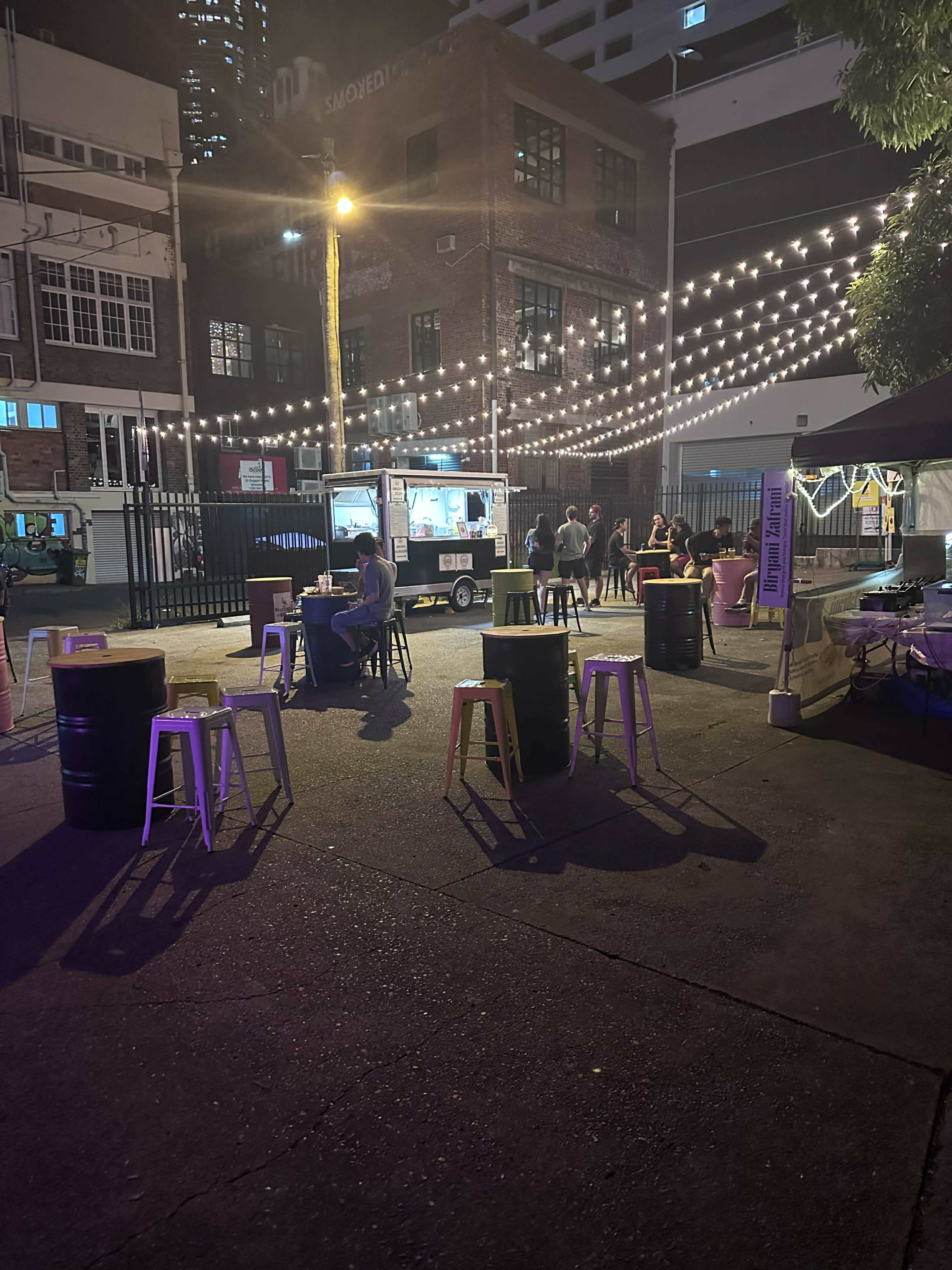 The scene shows an outdoor area with string lights overhead, several tables and stools, a food cart, and people socializing in the background.