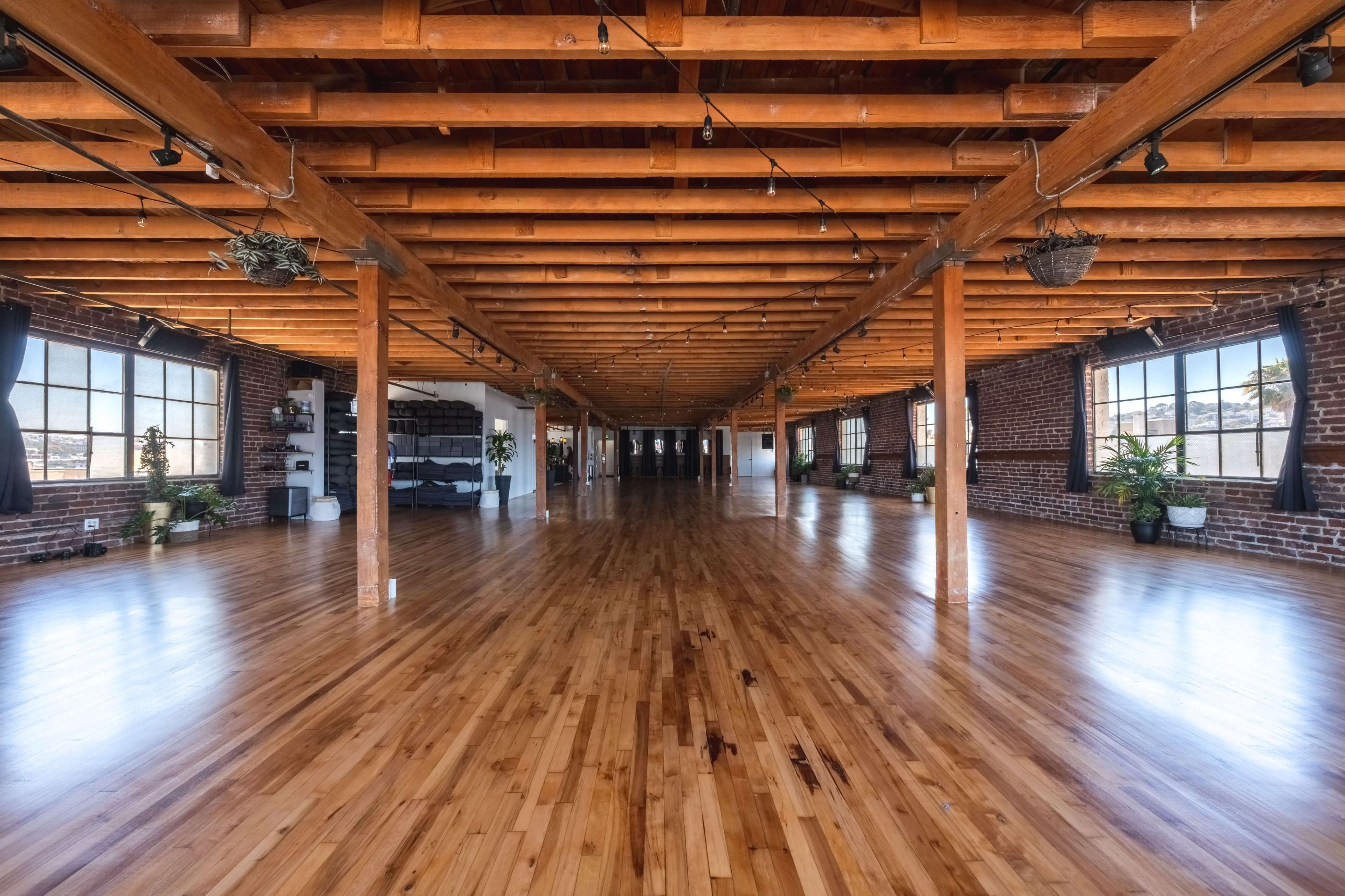 A spacious room with wooden beams and a polished hardwood floor features large windows and potted plants along the walls.