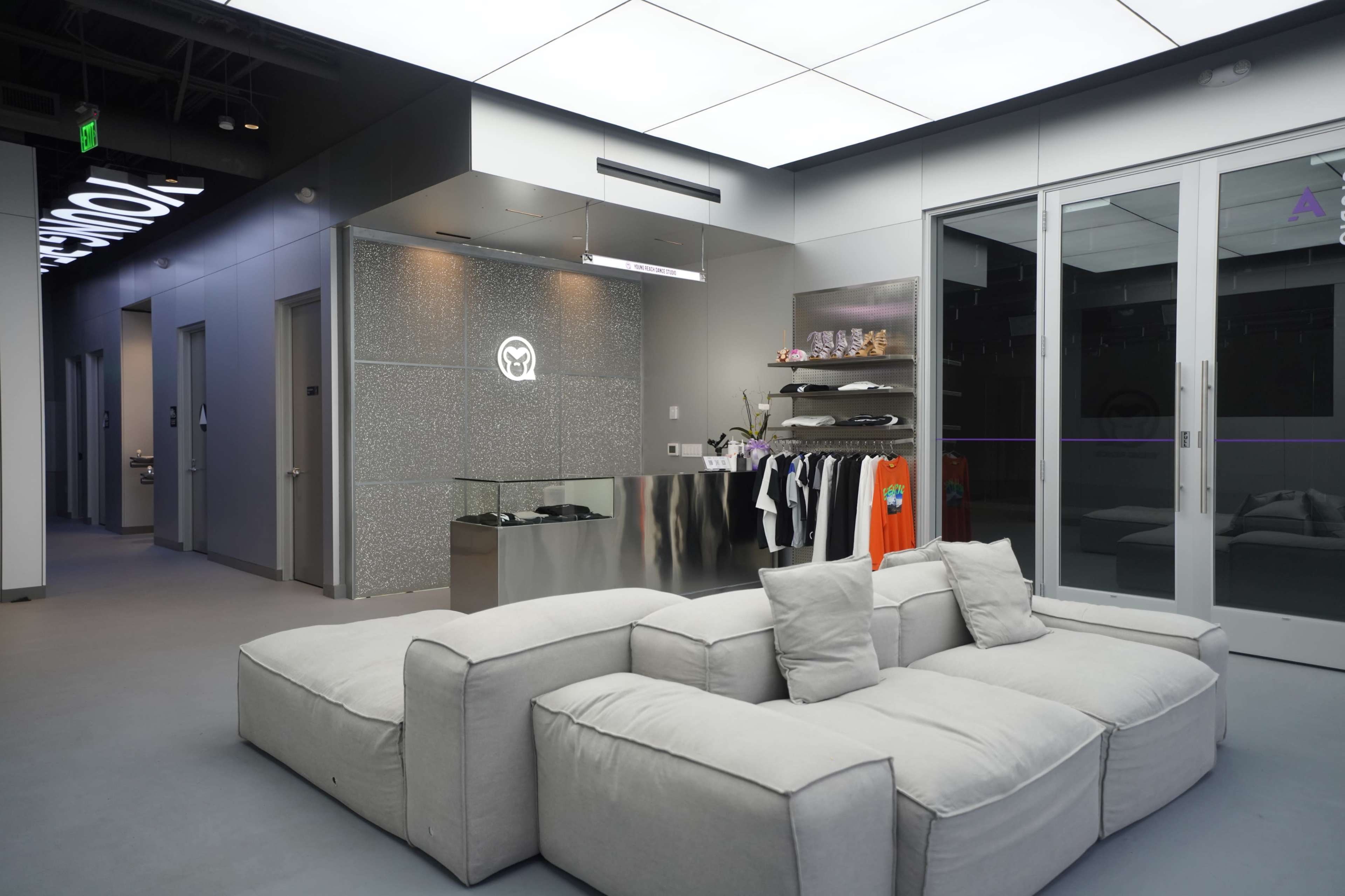 The image shows a modern interior space with a light-colored couch, a reception area featuring a logo, and clothing displayed on shelves against the wall.