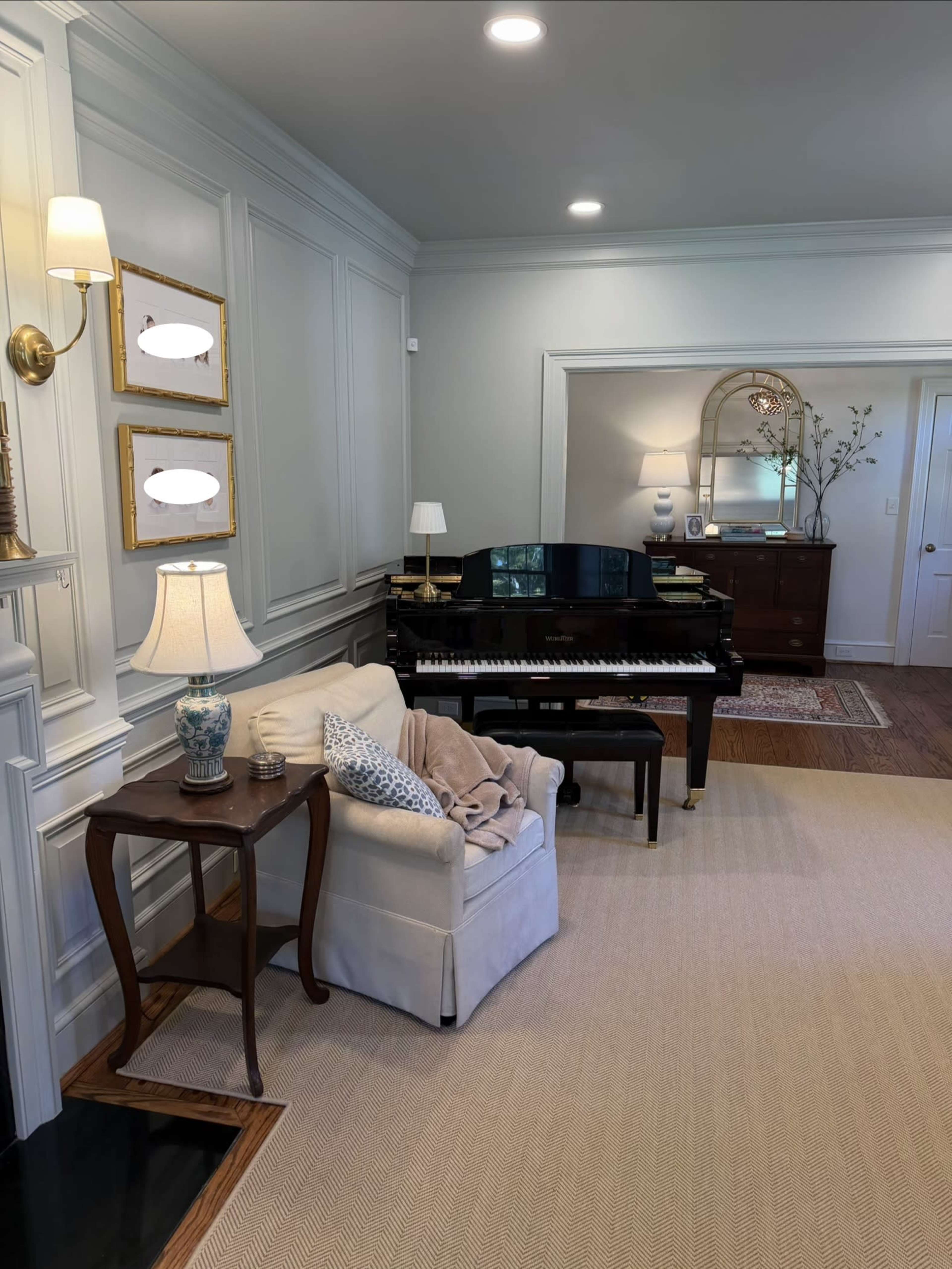 The image shows a well-lit room featuring a piano, a small table with a lamp, a cozy chair with a blanket, and decorative elements on the walls.