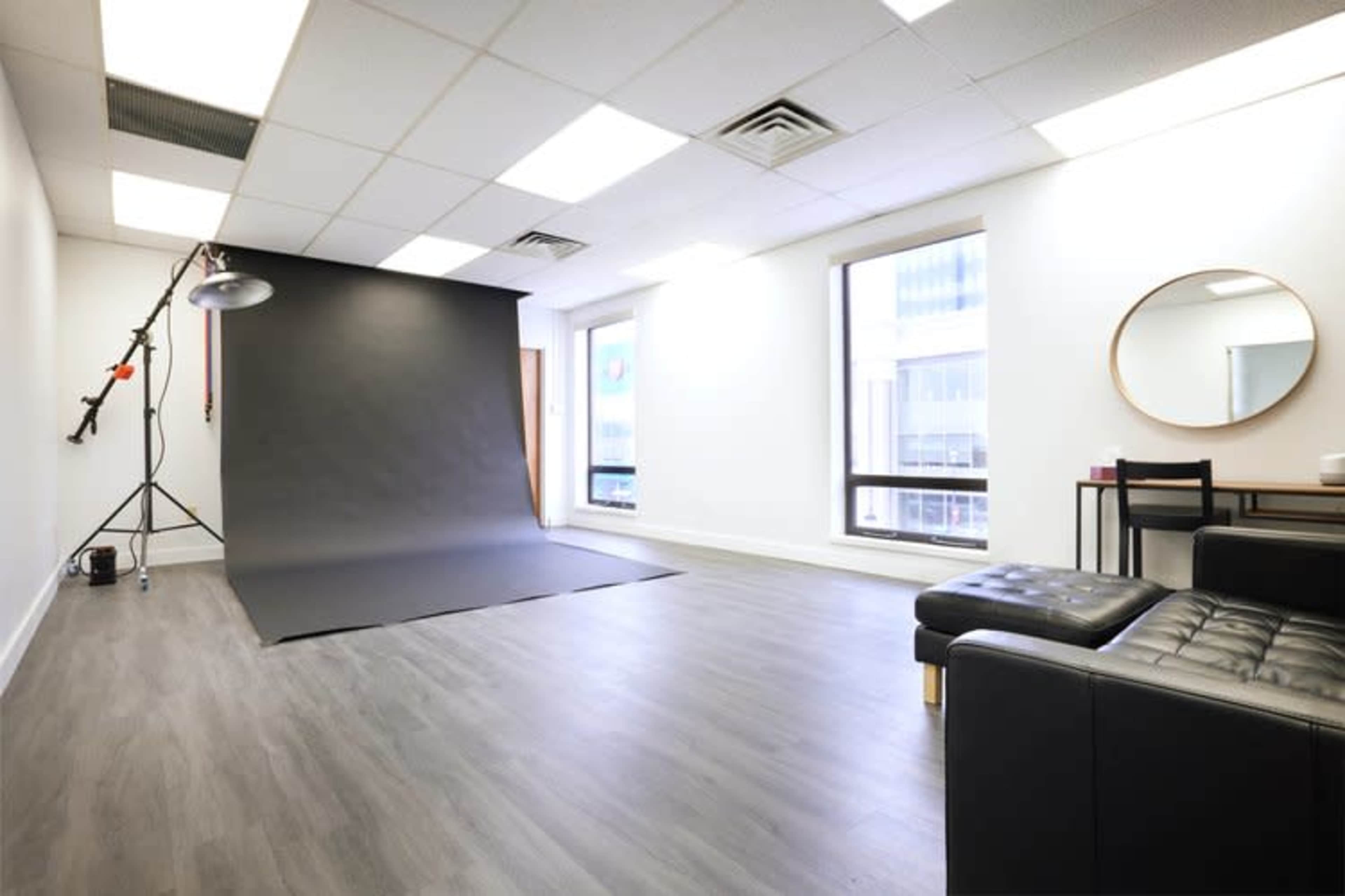 A photography studio with a black backdrop, a light stand, and a modern seating area, featuring large windows that let in natural light.
