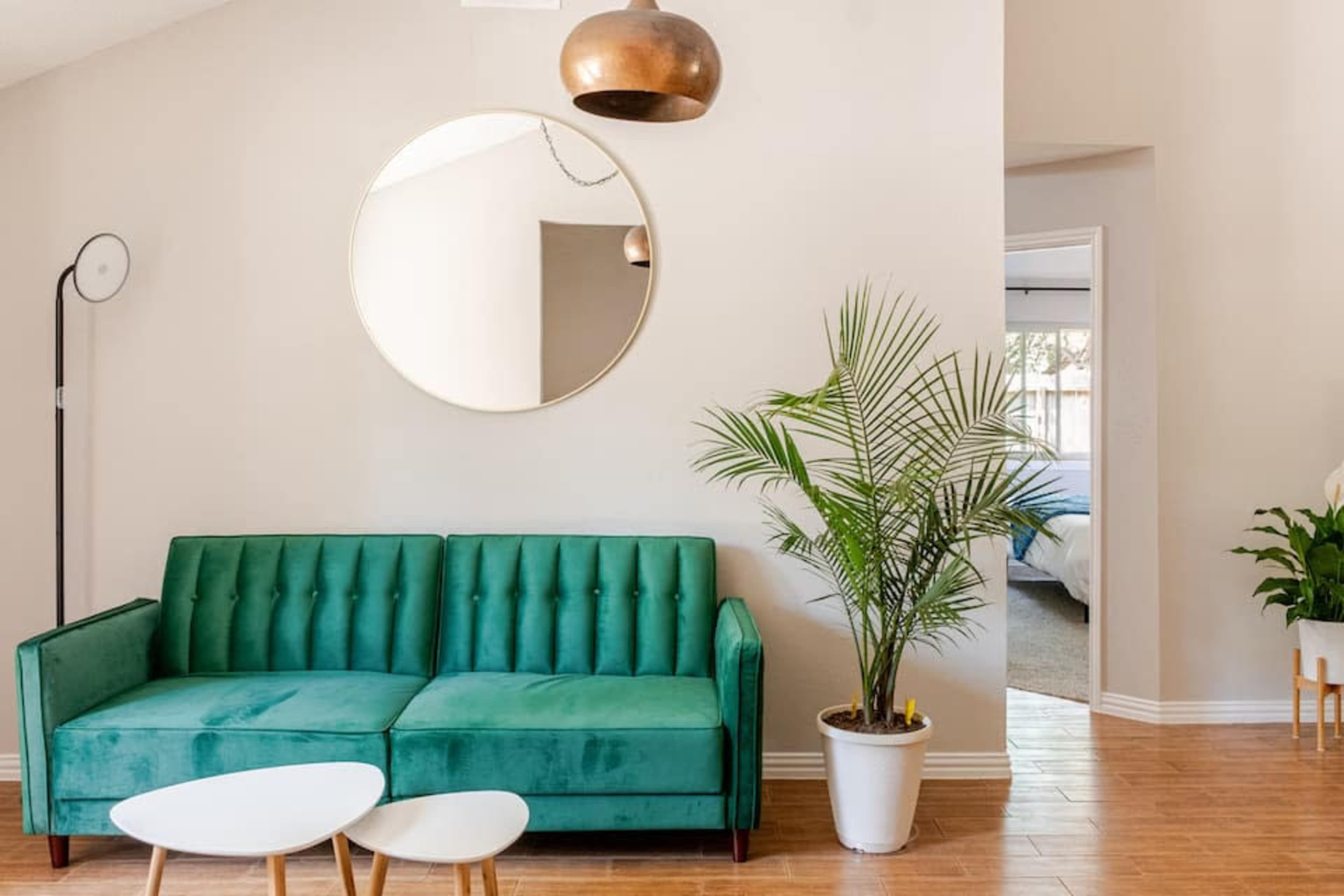 A green tufted sofa sits against a light wall, accompanied by a round mirror, a small table, and a potted plant.