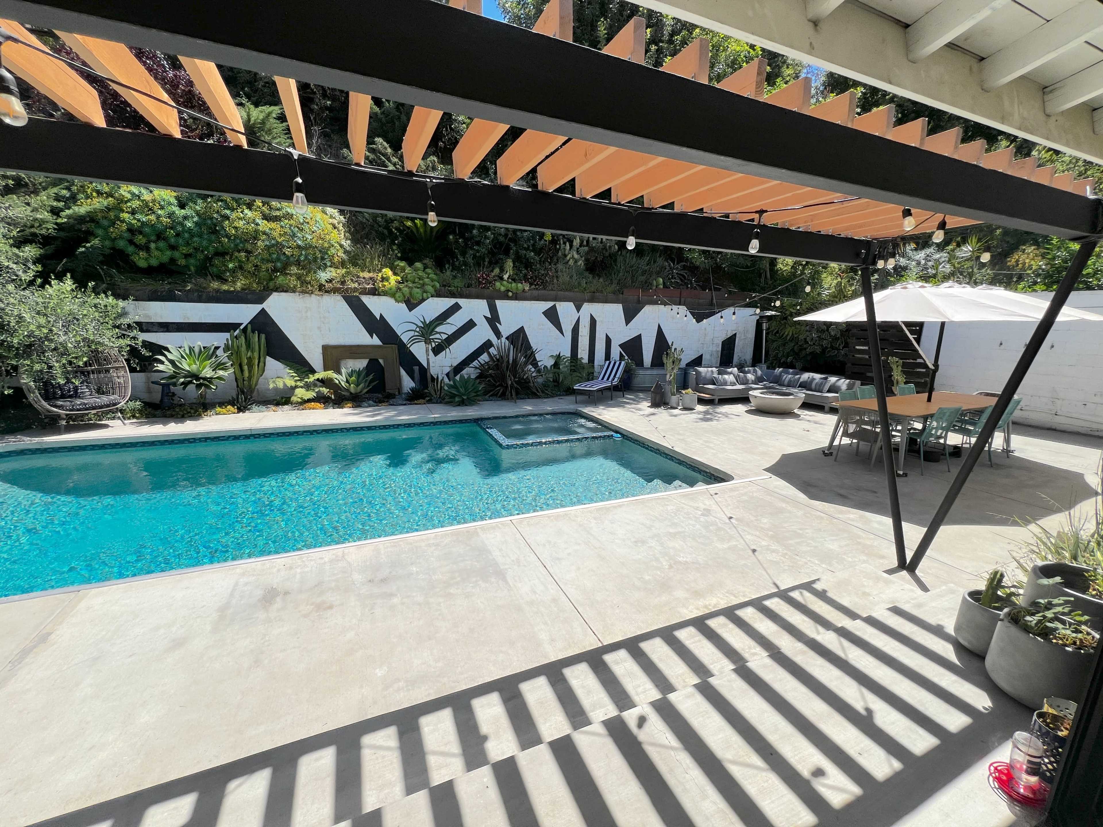 The image shows a backyard with a swimming pool, surrounded by plants and outdoor furniture, under a pergola with decorative overhead slats.