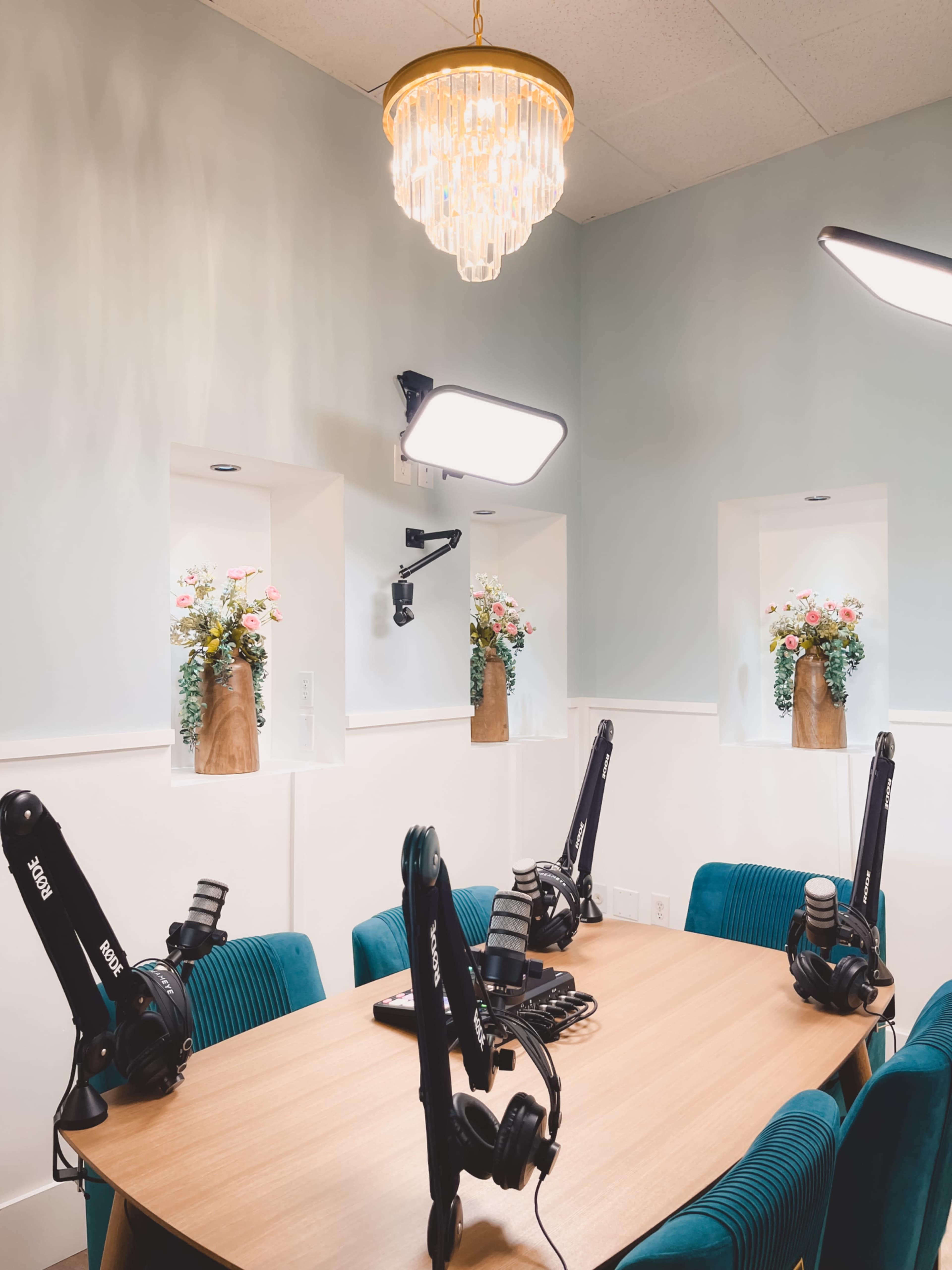 A podcast recording studio is set up with a wooden table, several microphones on adjustable arms, and decorative flower arrangements in wall niches.