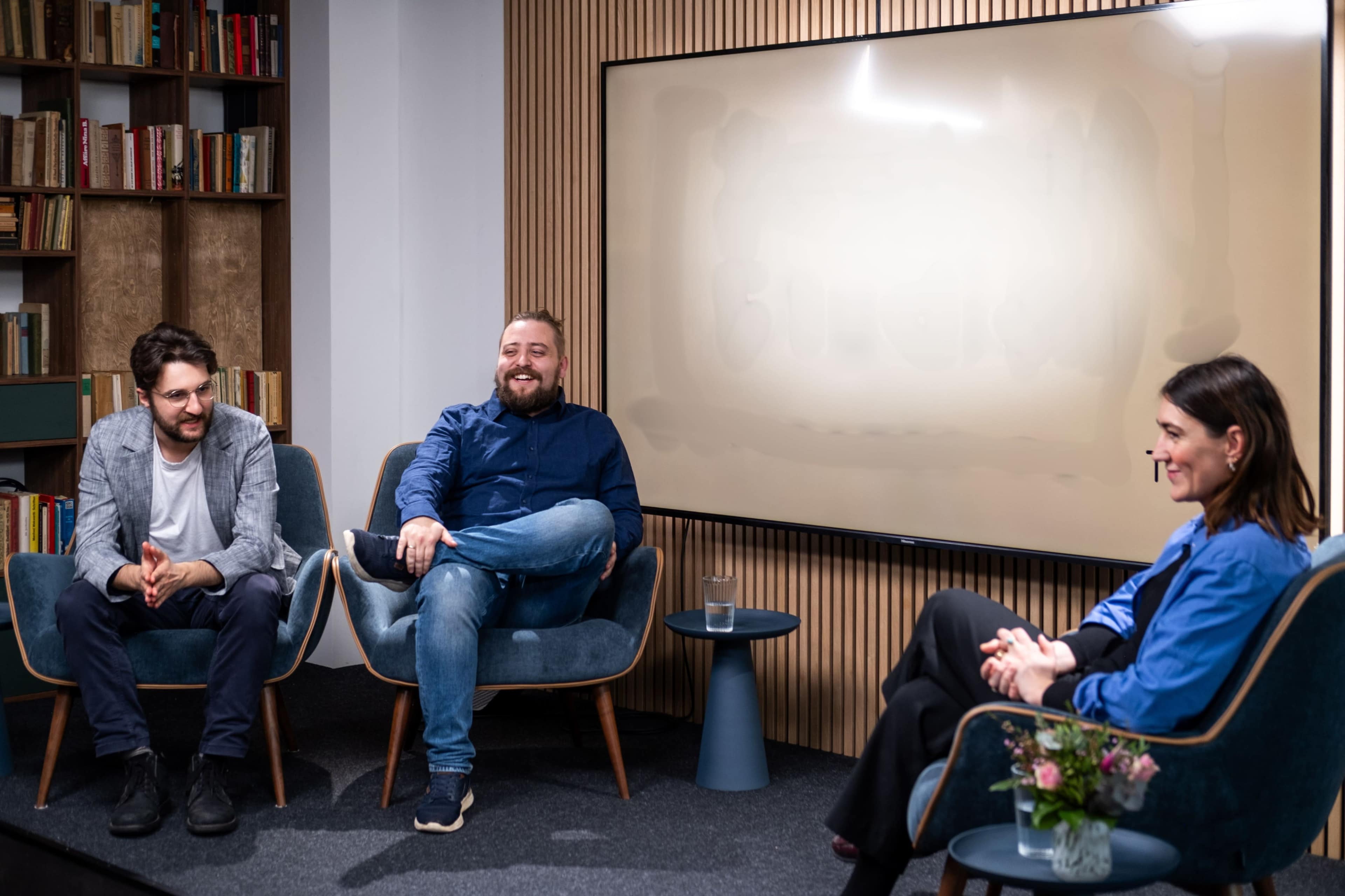 Three individuals are seated in modern chairs in a well-lit room decorated with bookshelves, engaging in conversation while facing a blank screen.