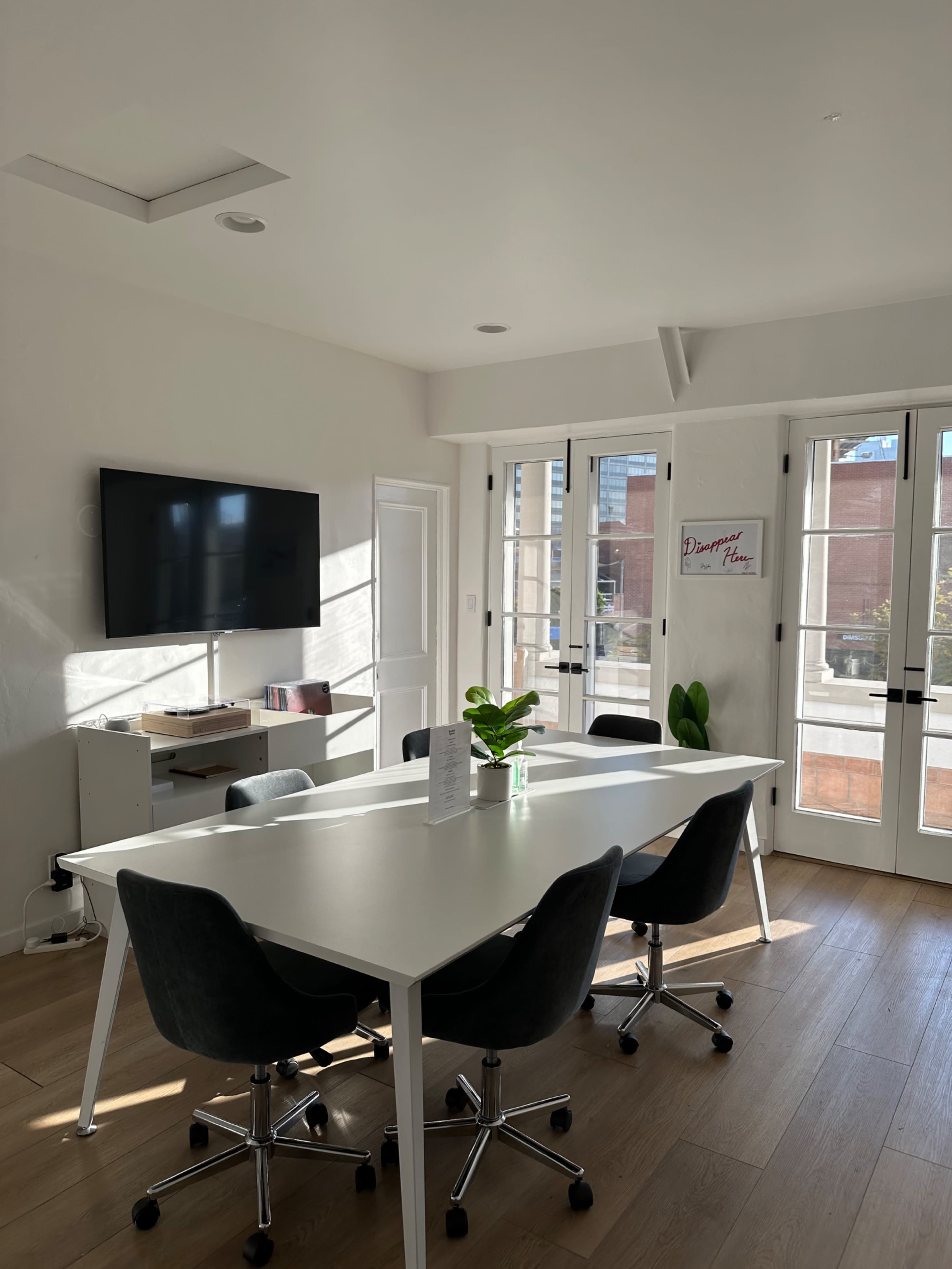 A modern conference room features a long white table surrounded by black rolling chairs, with a large TV mounted on the wall and windows providing natural light.