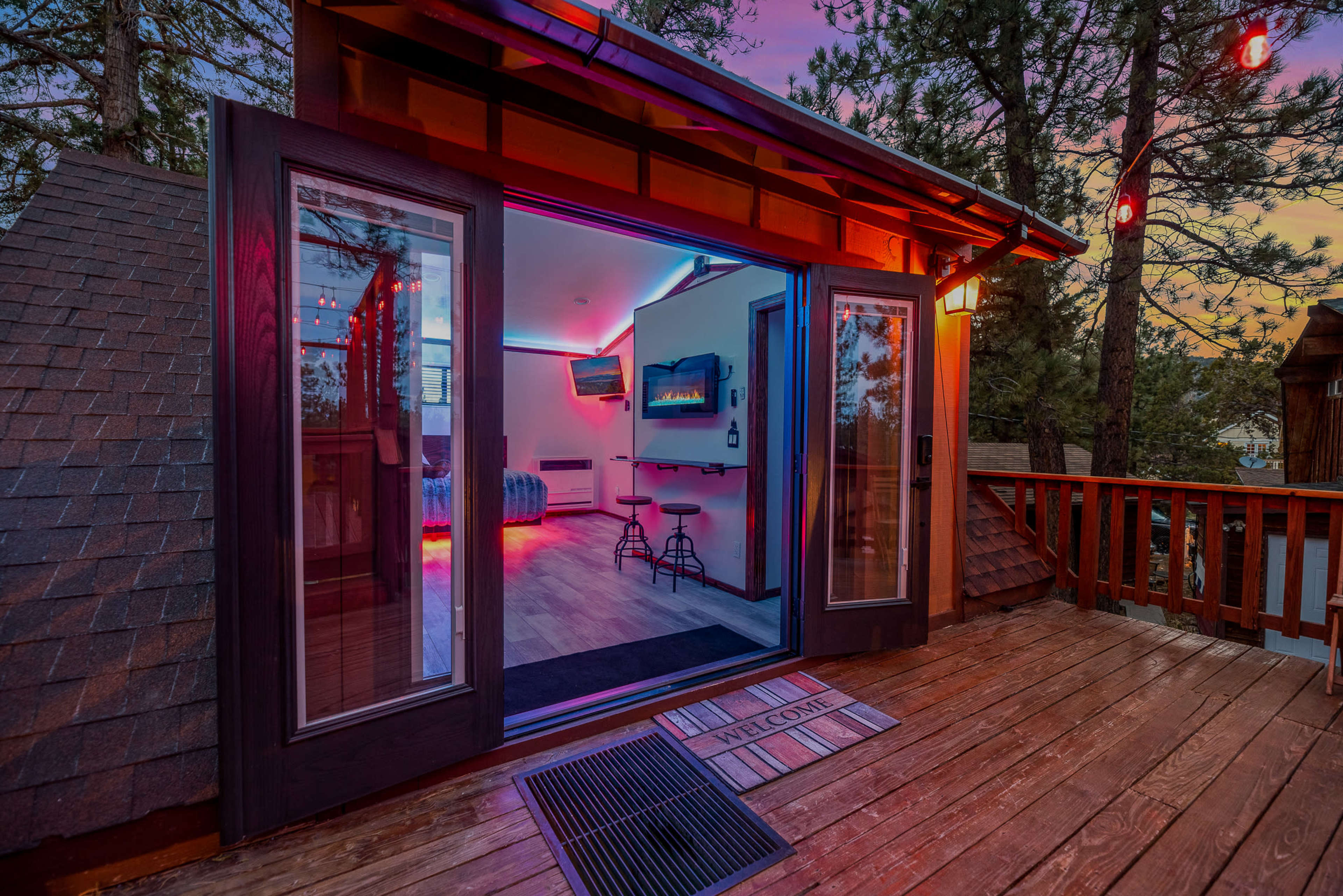 A modern cabin features large sliding glass doors opening onto a wooden deck, surrounded by tall trees and illuminated by colored lights.