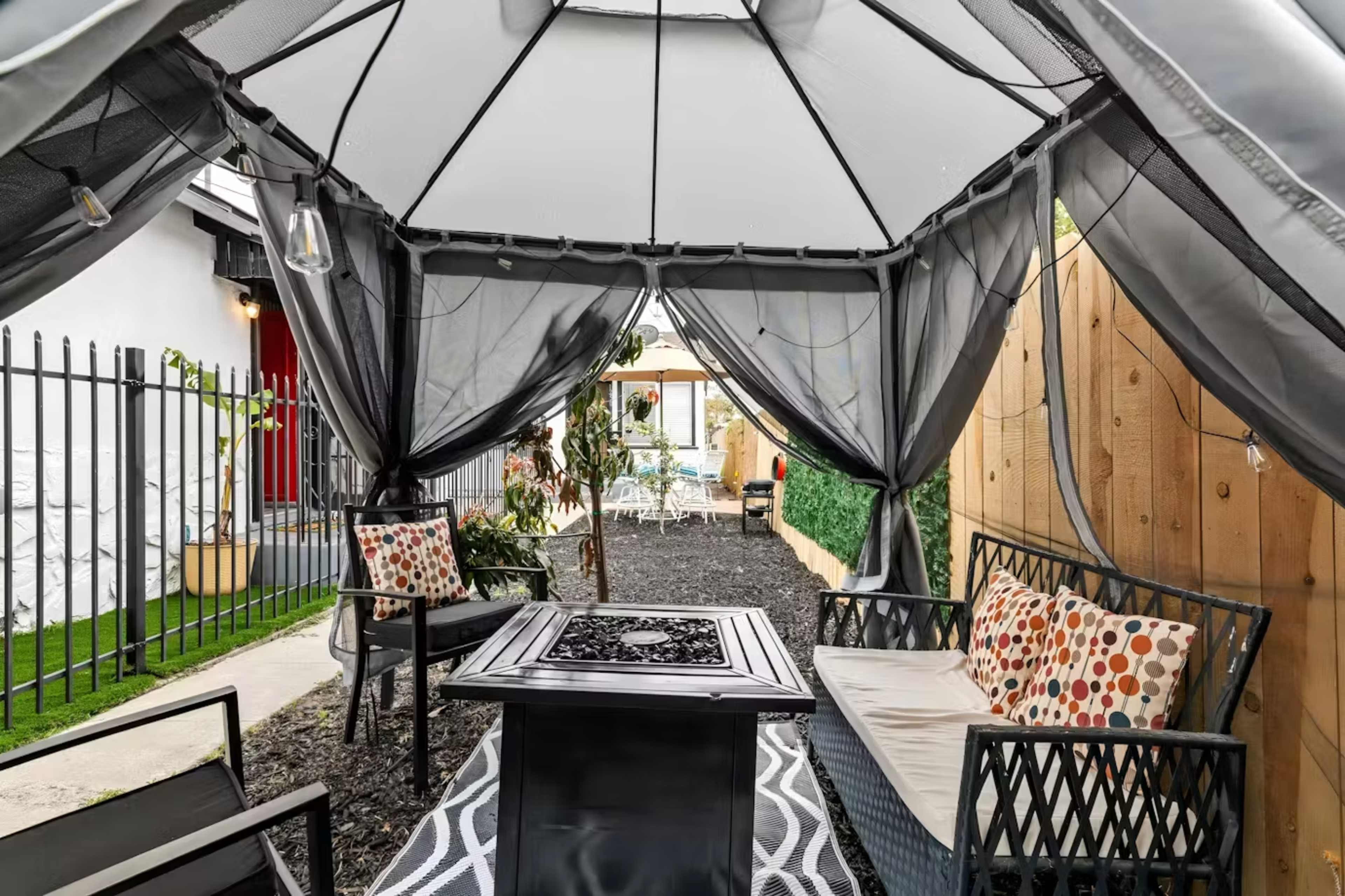 A cozy outdoor sitting area features a gazebo with draped curtains, two patterned chairs, and a central fire table surrounded by gravel.