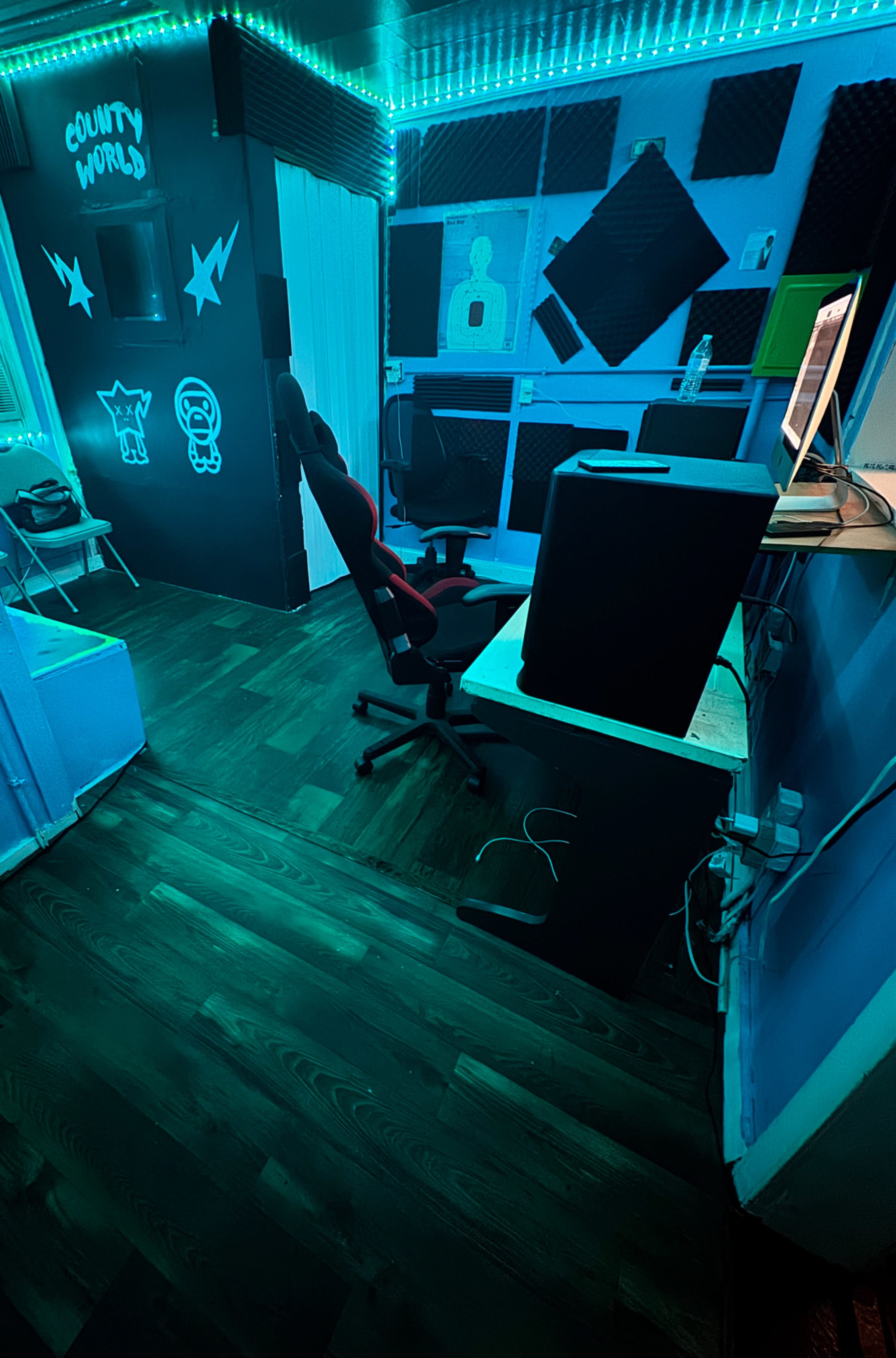 The image shows a dimly lit recording studio with a black chair, a computer on a desk, and soundproofing panels on the walls.