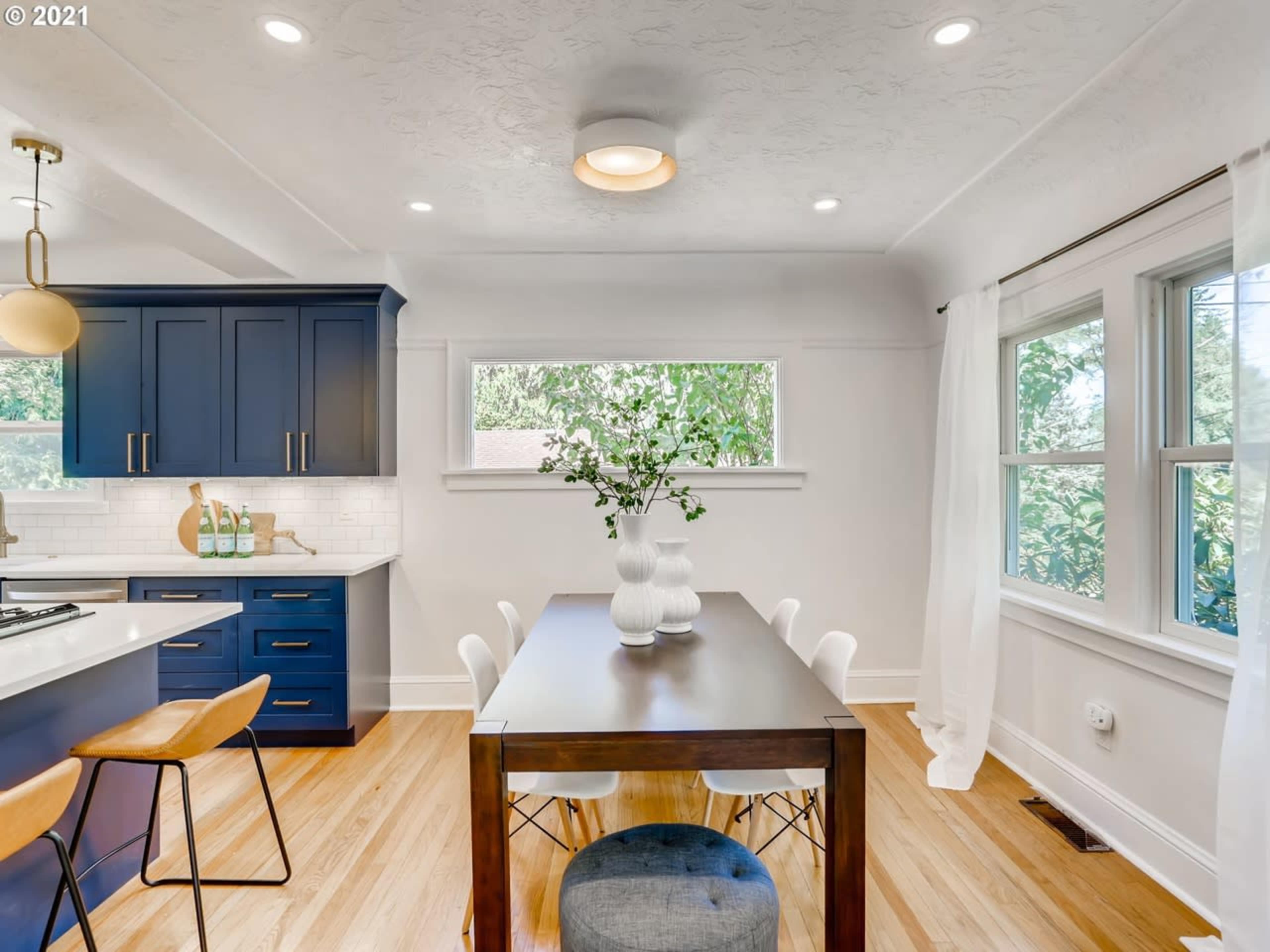 A modern kitchen and dining area features blue cabinetry, a wooden dining table, and large windows allowing natural light.