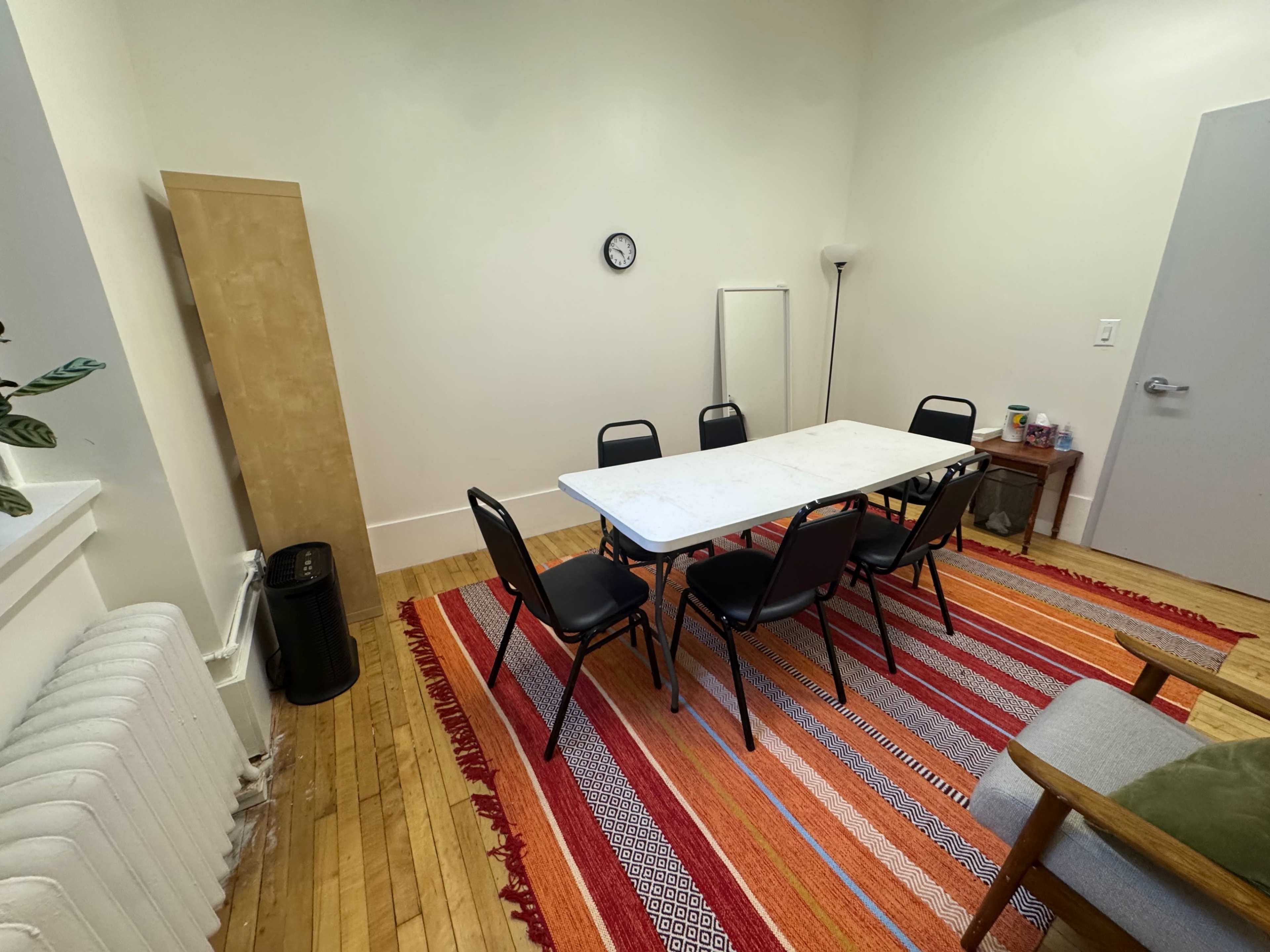 The image shows a simple meeting room with a rectangular table surrounded by black chairs, a clock on the wall, and a rug on the wooden floor.