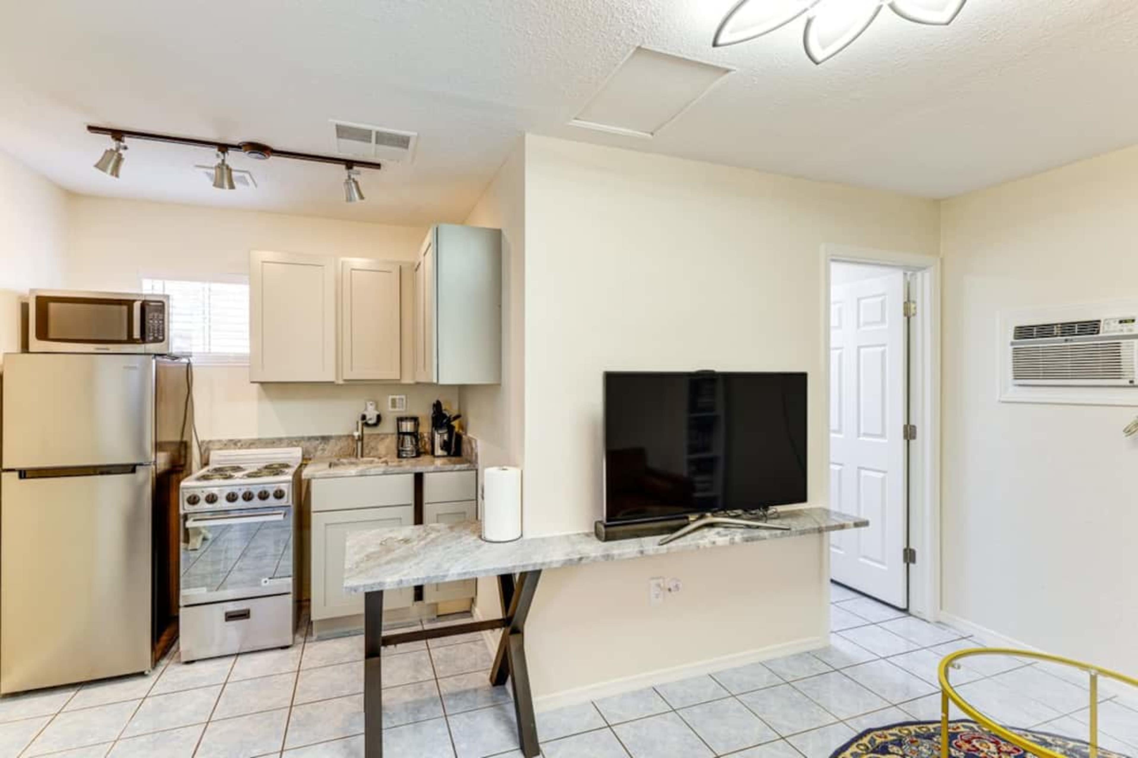 The image shows a small kitchen area with a refrigerator, stove, coffee maker, and a television on a table, next to a door and an air conditioning unit.