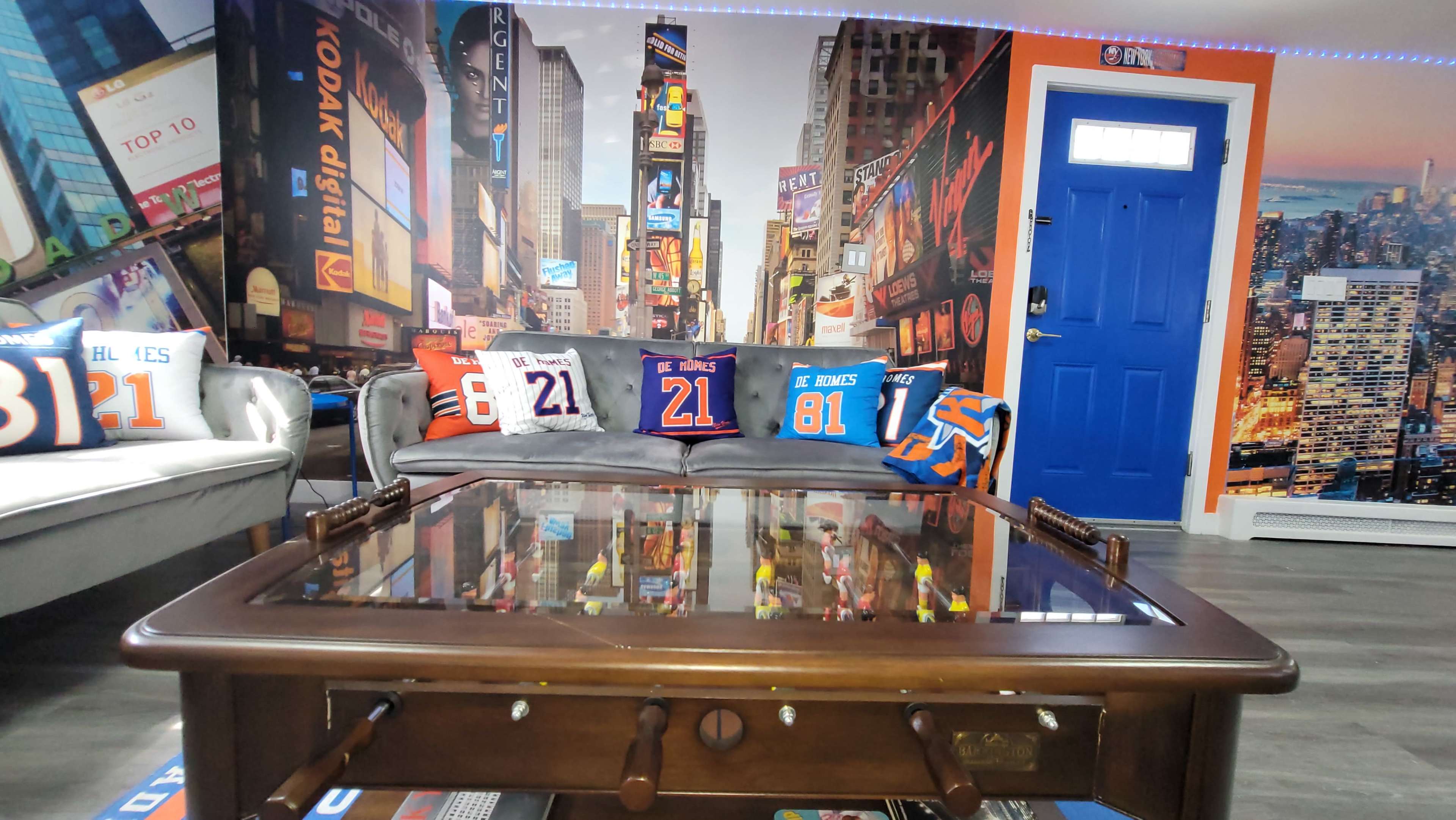 A brightly decorated room featuring a couch with sports-themed throw pillows, a wooden coffee table with a glass top, and a mural of a vibrant cityscape on the walls.