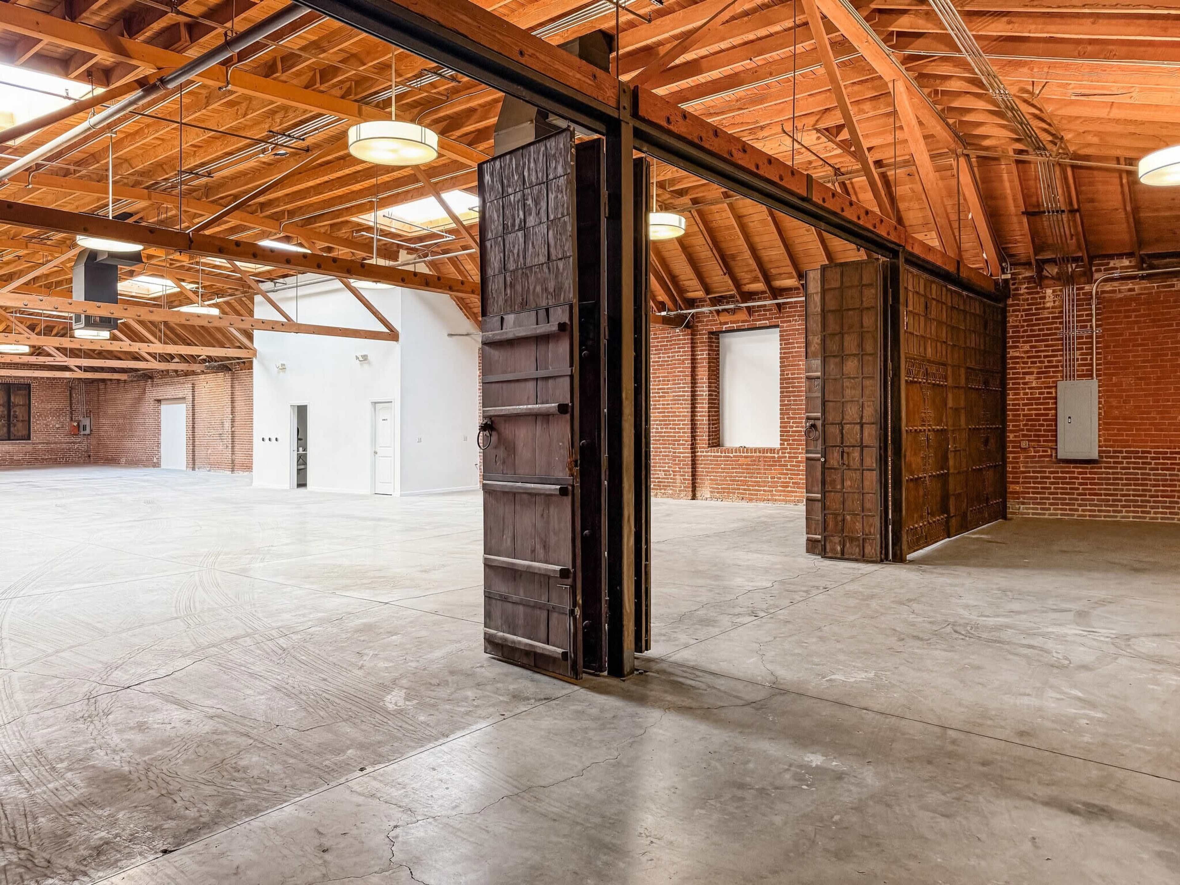 The image shows a spacious industrial interior with exposed wooden beams, brick walls, and large sliding doors.
