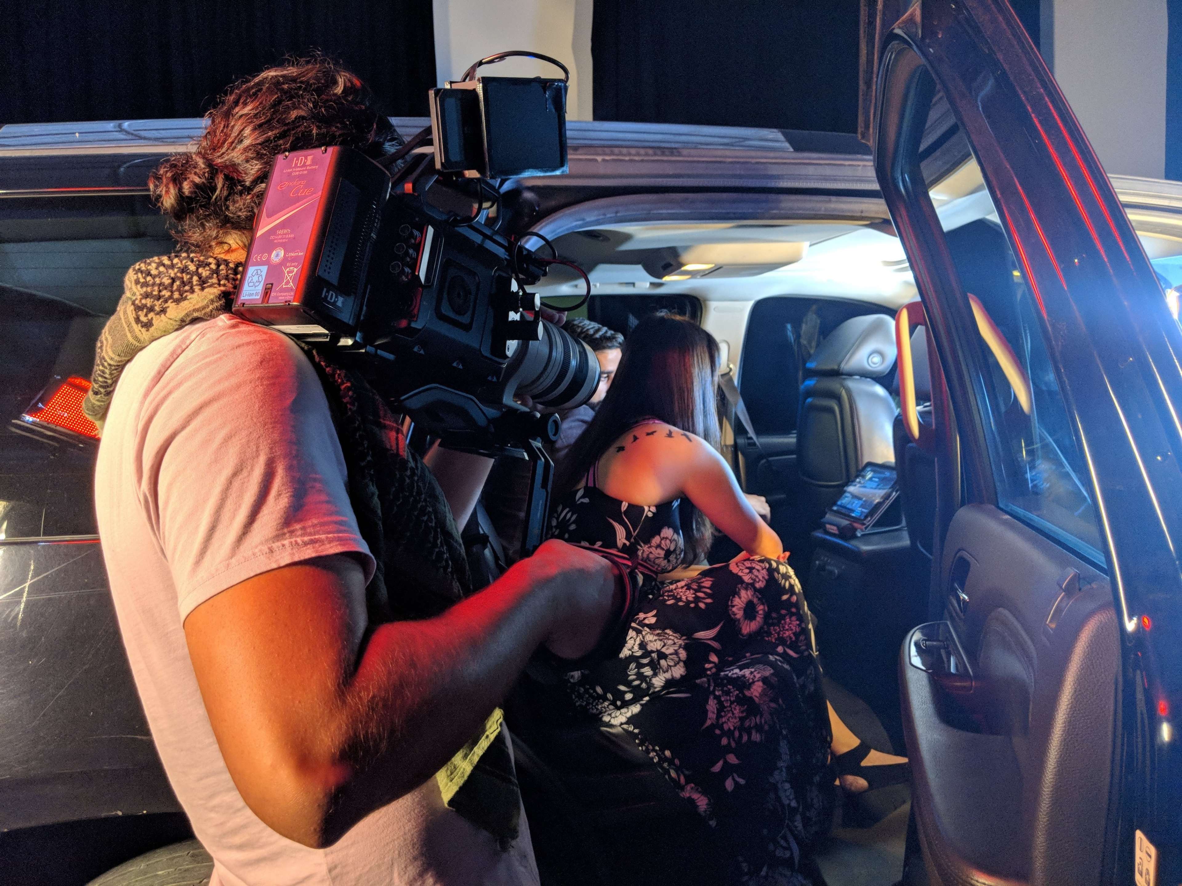 A camera operator films a woman sitting inside a vehicle, reviewing content on a device.