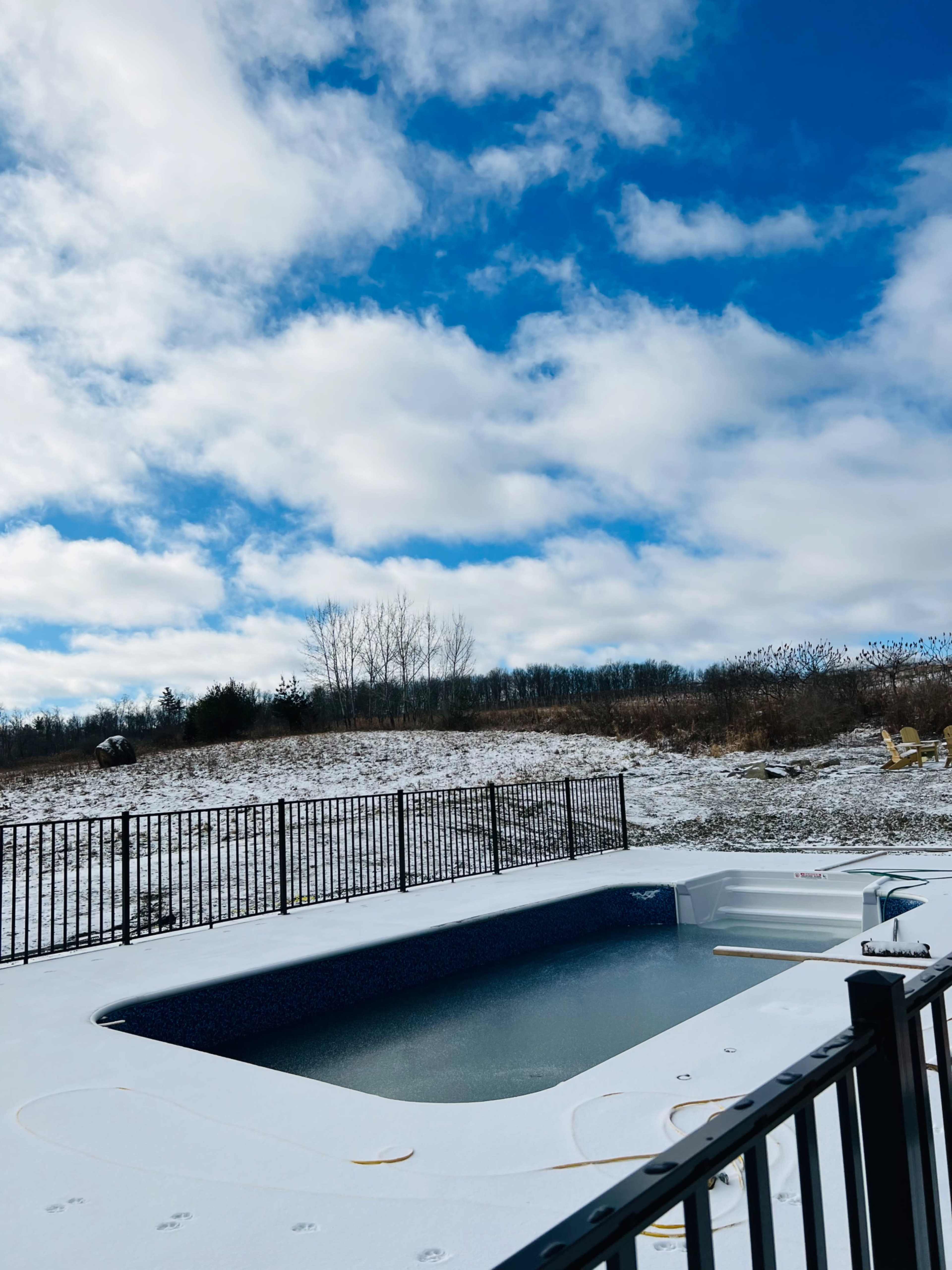 A snow-covered pool area is surrounded by a black fence under a partly cloudy sky.