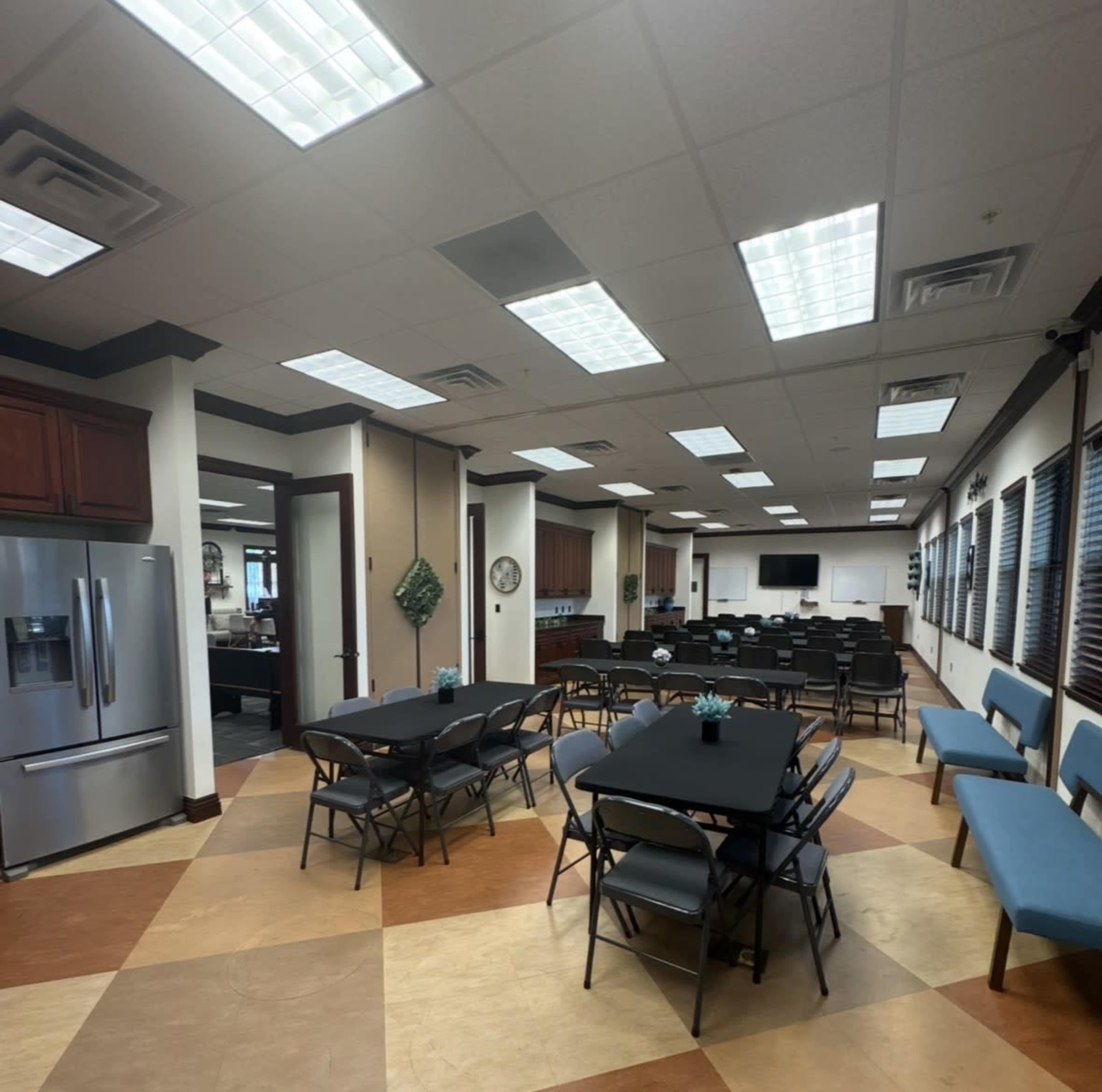 The image shows a spacious meeting room with several tables and chairs arranged neatly, a kitchenette area, and a large screen at the far end.